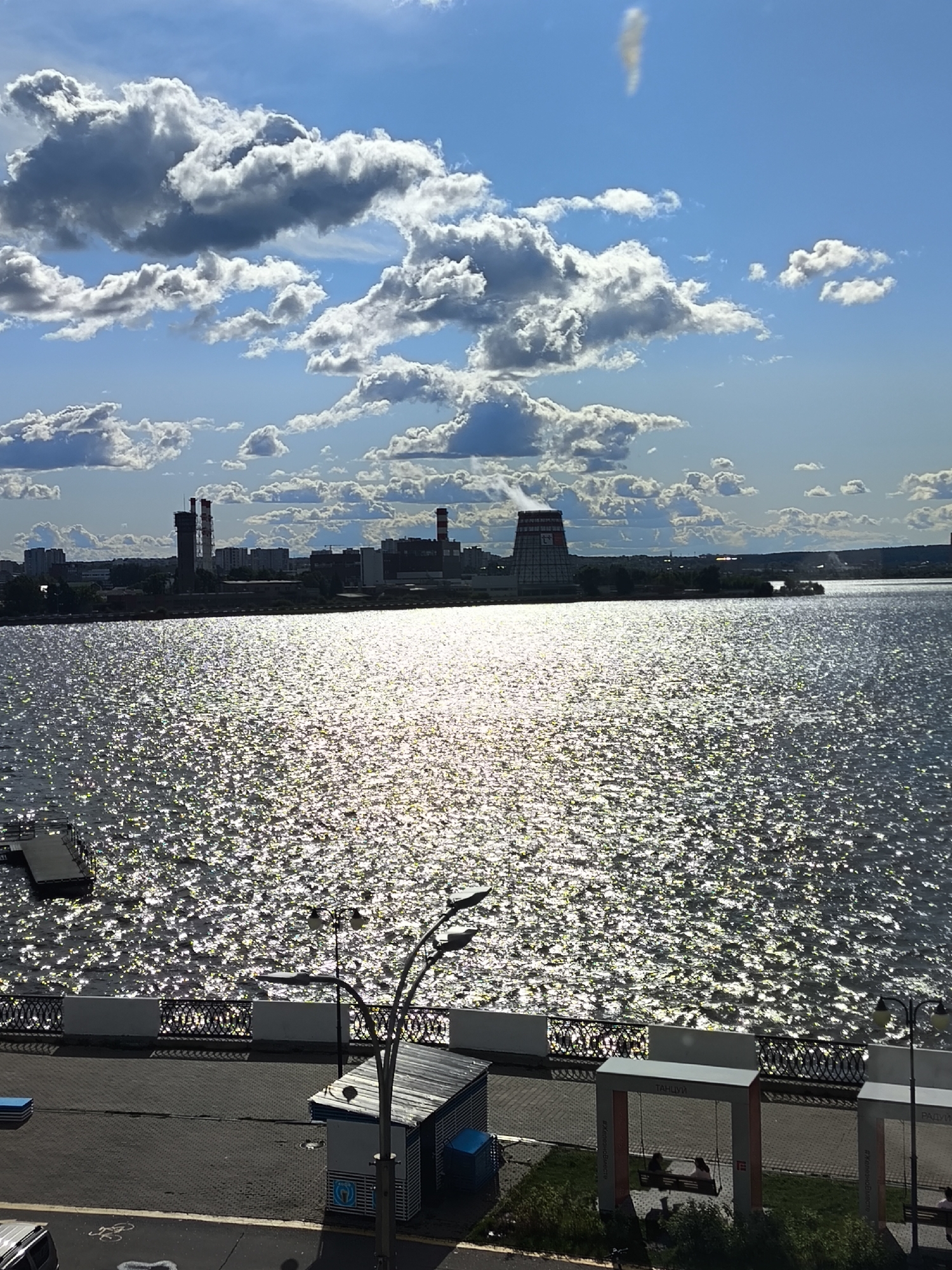 This image captures a scenic view of the Izhevsk Reservoir in Izhevsk, Russia, on a bright, partly cloudy day. The sun glints off the water's surface, creating a shimmering effect. In the distance, the city skyline is visible, featuring industrial structures like chimneys and buildings. The foreground shows a paved walkway along the waterfront, with a decorative fence. There is a small pier extending into the water on the left. On the right, a modern structure with swings is present, and two individuals are seated on one of them. There are also lampposts and a small kiosk-like building. The overall scene depicts a recreational area by the water, with the urban landscape as a backdrop.
