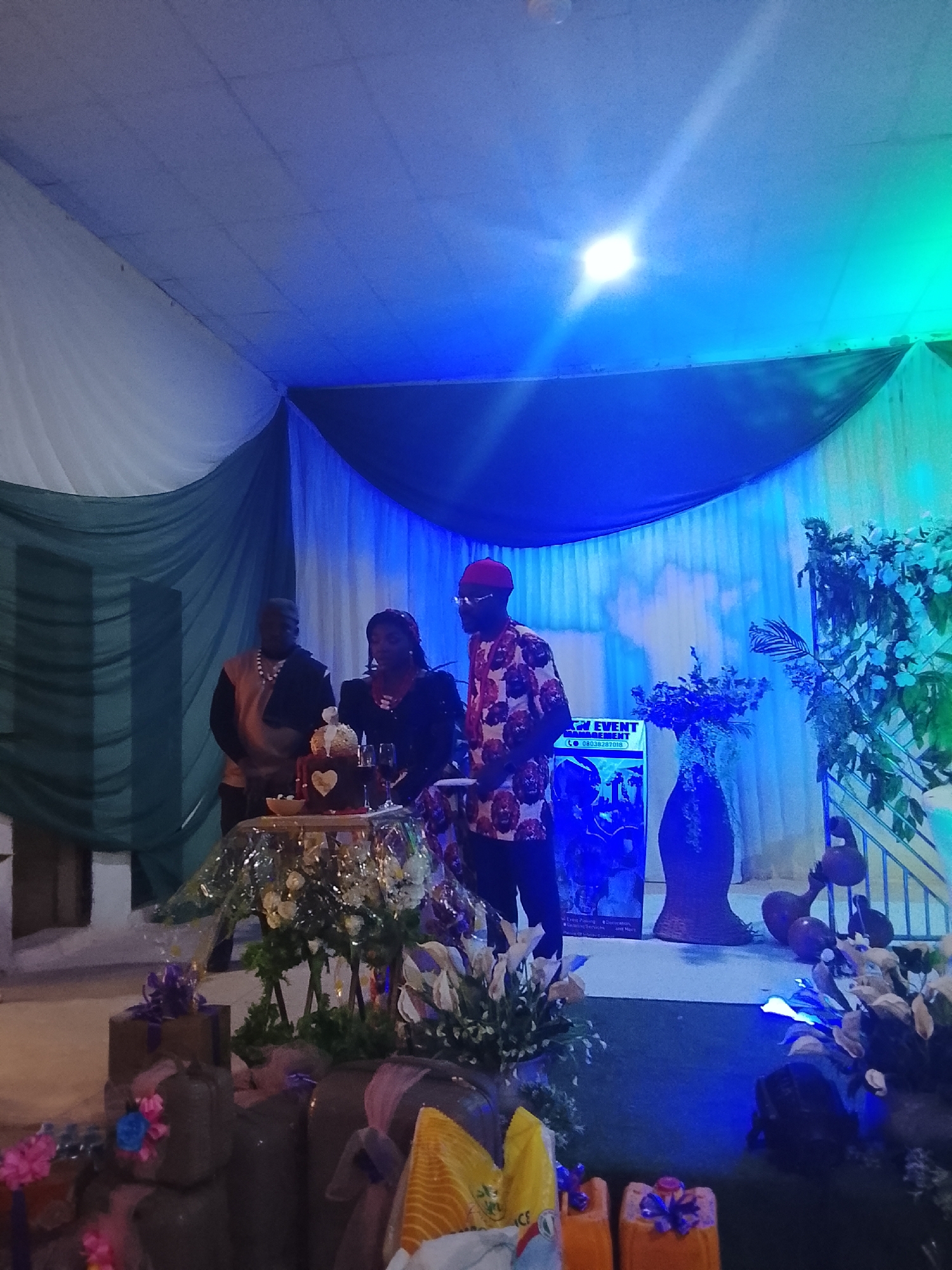The image depicts a wedding reception held in a hall in Vom, Nigeria. Three people are standing behind a table holding a wedding cake. A man on the left wears a dark traditional outfit. A woman in the middle wears a dark dress and necklace. A man on the right wears a patterned shirt and red cap. The table is decorated with white flowers and a silver cloth. Gifts, decorations, and vases of flowers are arranged around the table. The hall is decorated with white and green curtains and illuminated by blue and green lights. A sign indicating an event management company is partially visible to the right.