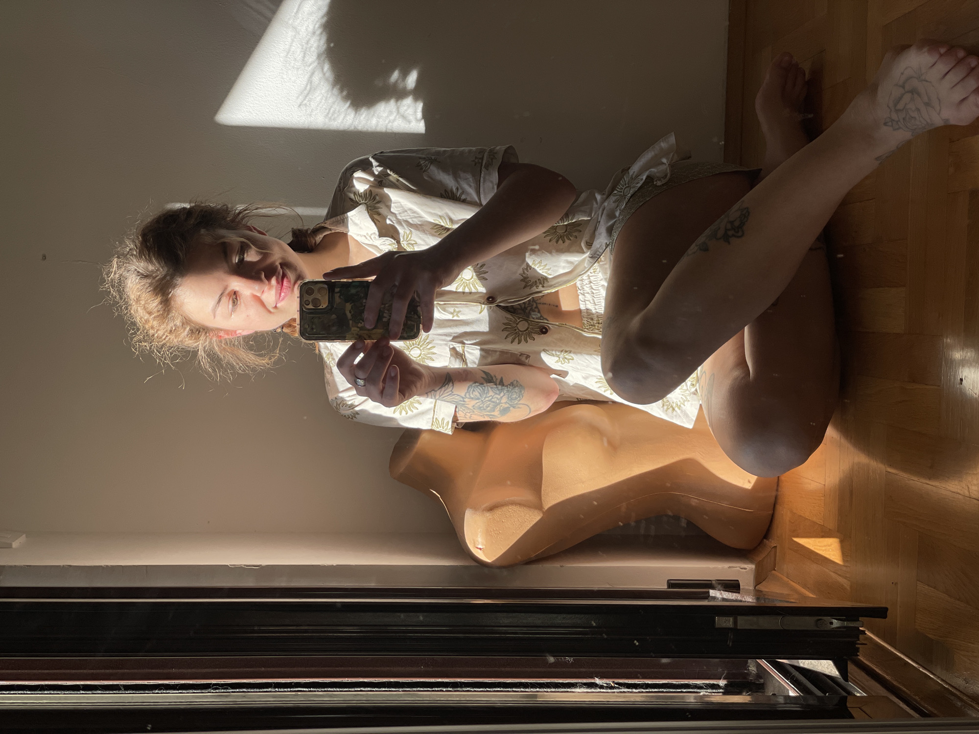 The image appears to be a selfie taken by a woman in an indoor setting. She is holding a smartphone with a camouflage-patterned case. The woman is fair-skinned with light brown hair and is wearing a light-colored, patterned shirt. Tattoos are visible on her left arm, leg, and foot. She is positioned next to a mannequin torso. The room has hardwood flooring and a wall with a window, partially obscured by blinds or a closed window frame. Sunlight casts shadows on the wall and floor. The location in the city of Boreti, Montenegro, is not evident from the image.