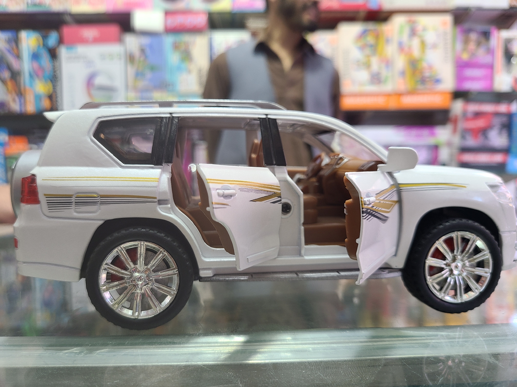 The image shows a white toy SUV with the front and rear doors open, revealing a brown interior. It's positioned on a reflective surface, possibly glass, with a blurry background that suggests a store setting, possibly in Quetta City Tehsil, Pakistan, given the request. In the background, there's a partially visible person with a beard wearing a vest. Behind the toy and the person is a display of colorful items, appearing to be books or similar products on shelves.