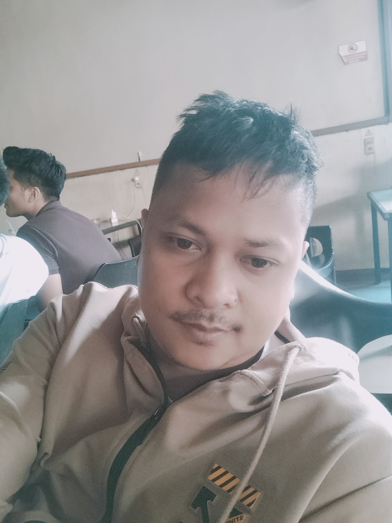 A close-up selfie shows a young man with short, dark, spiky hair and a mustache. He is wearing a light-colored hooded sweatshirt with a dark zipper. To his left, another young man with short dark hair is visible, wearing a dark t-shirt and looking away from the camera. The background is blurred, but suggests an indoor setting with neutral-colored walls, a wooden trim, and some furniture. The lighting appears to be indoor. The specific location in Meulaboh, Indonesia, is not identifiable from the visual information in the image.
