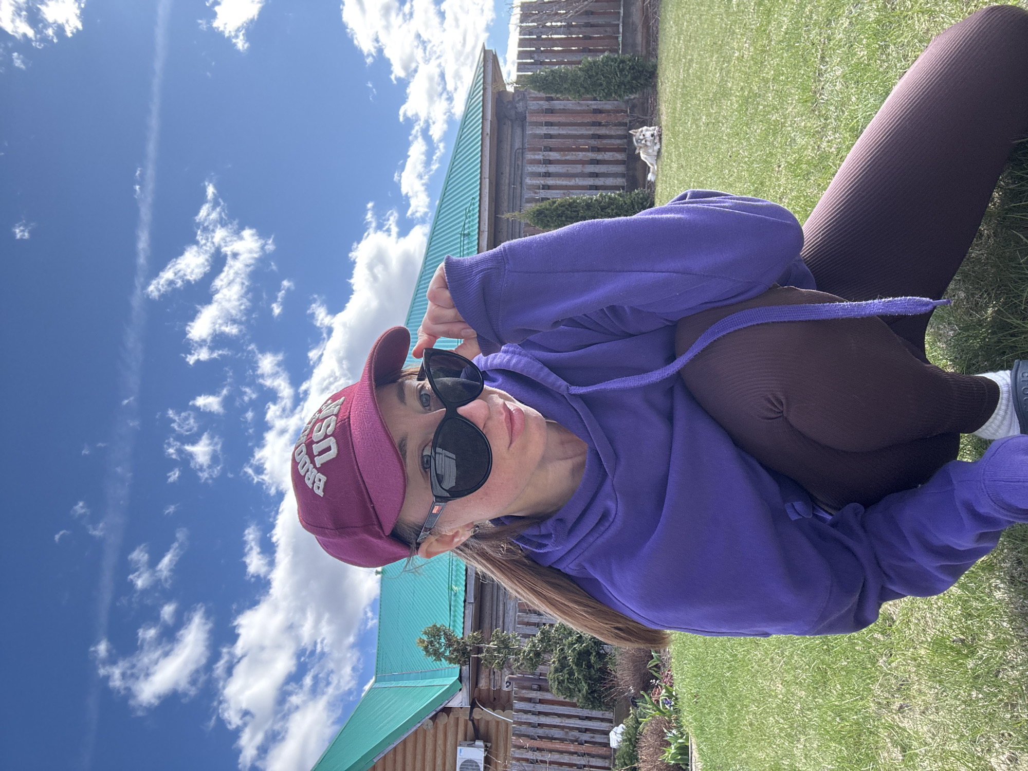 A woman in a purple hoodie and maroon leggings sits on grass outdoors. She wears a maroon baseball cap with "BROOK USA" on the front and black sunglasses, which she is adjusting with her right hand. Behind her, a wooden building with a green roof stands under a blue sky with scattered white clouds. A wooden fence and evergreen trees are visible behind the building. A white dog stands near the fence. A split air conditioner is mounted on the wooden building, suggesting an indoor temperature control feature. The location is presumably near the city of Прикамский, Russia, based on the original query.