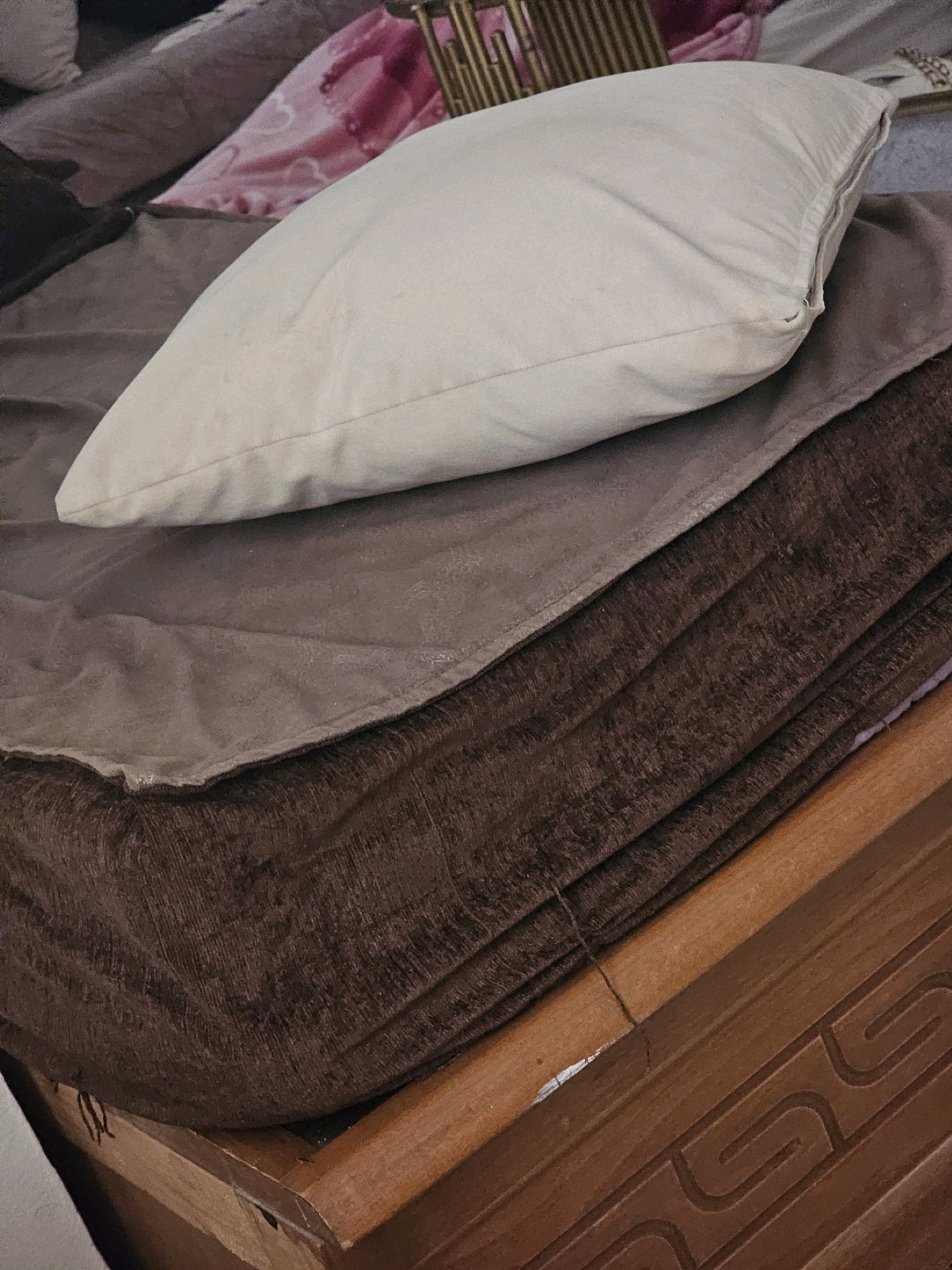 The image shows a close-up of a stack of folded blankets and a pillow on a bed. The blankets are in various shades of brown and taupe, with one blanket appearing to have a textured, ribbed surface. A light-colored pillow rests on top of the folded blankets. In the background, a pink blanket with a heart pattern is visible, along with a portion of a gold-colored decorative stand. The bed frame, made of dark wood with carved detailing, is visible at the bottom of the image. The scene appears to be indoors, likely in a bedroom. There is no indication of the city of Batna, Algeria, or any specific location within it.