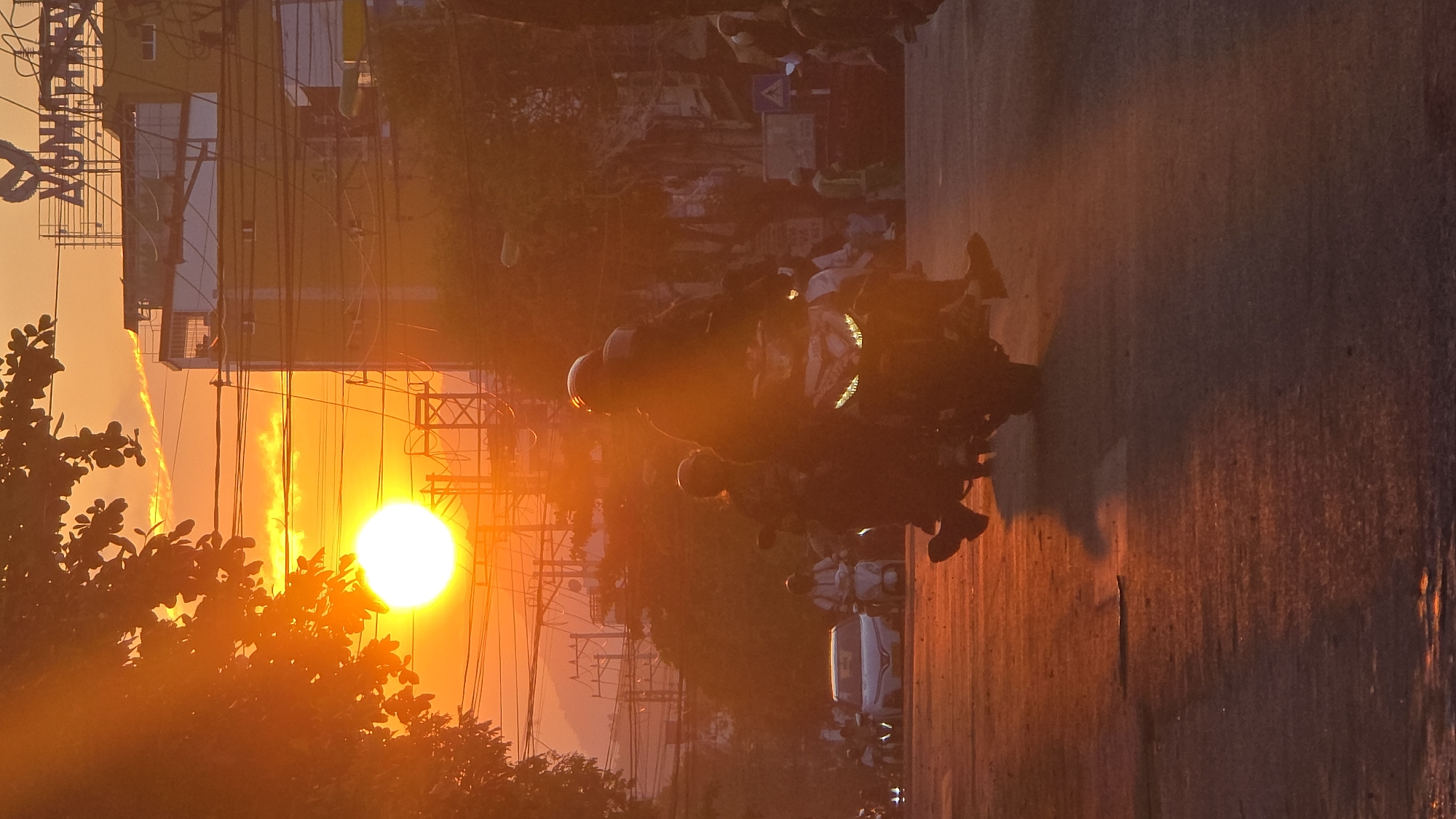 The image captures an urban street scene in Nha Trang, Vietnam, during either sunrise or sunset. A large, brilliant orange sun is prominently positioned in the upper left quadrant, casting an intense, monochromatic orange-red light across the entire scene and creating strong backlighting.
In the foreground, a wet or reflective asphalt road surface is visible. A dark-colored motor scooter occupies the mid-foreground, traveling away from the viewer. It is ridden by two individuals, both wearing helmets. The scooter's LED headlights are illuminated. A second motor scooter, also carrying two helmeted individuals, is visible to the left of the primary scooter. Further down the road, multiple additional motor scooters and at least one light-colored sedan are discernible as dark silhouettes. On the right side of the road, a pedestrian or worker wearing a light-colored, possibly high-visibility vest, is faintly visible.
The left side of the frame is partially obscured by silhouetted tree branches and foliage. On the right, a multi-story building with a yellow facade and reflective windows dominates the midground. A rooftop sign, featuring a stylized logo and partially legible text, is affixed to this structure. A dense array of overhead utility wires and cables crisscrosses the sky above the street, particularly in front of the building. In the distant background, silhouetted mountains or hills are present beneath a hazy sky. The scene depicts active urban traffic in highly dramatic natural lighting.