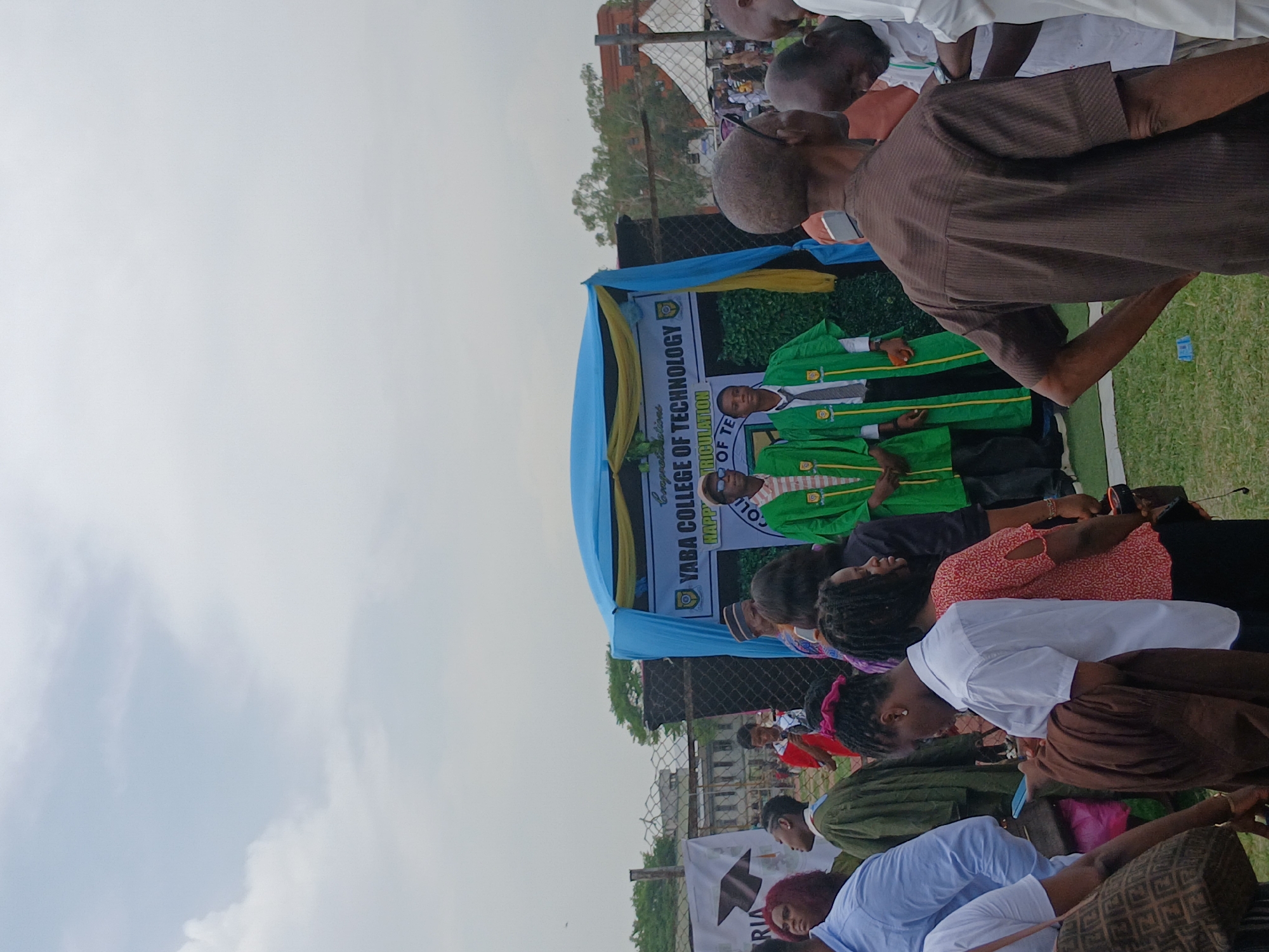 The image shows an outdoor event, possibly a graduation or matriculation ceremony, at Yaba College of Technology in Lagos, Nigeria. A banner with the college's name is visible, and people are standing on a stage, some wearing green academic gowns. An audience of people are seated in front of the stage. The sky is overcast, and a chain-link fence is in the background.