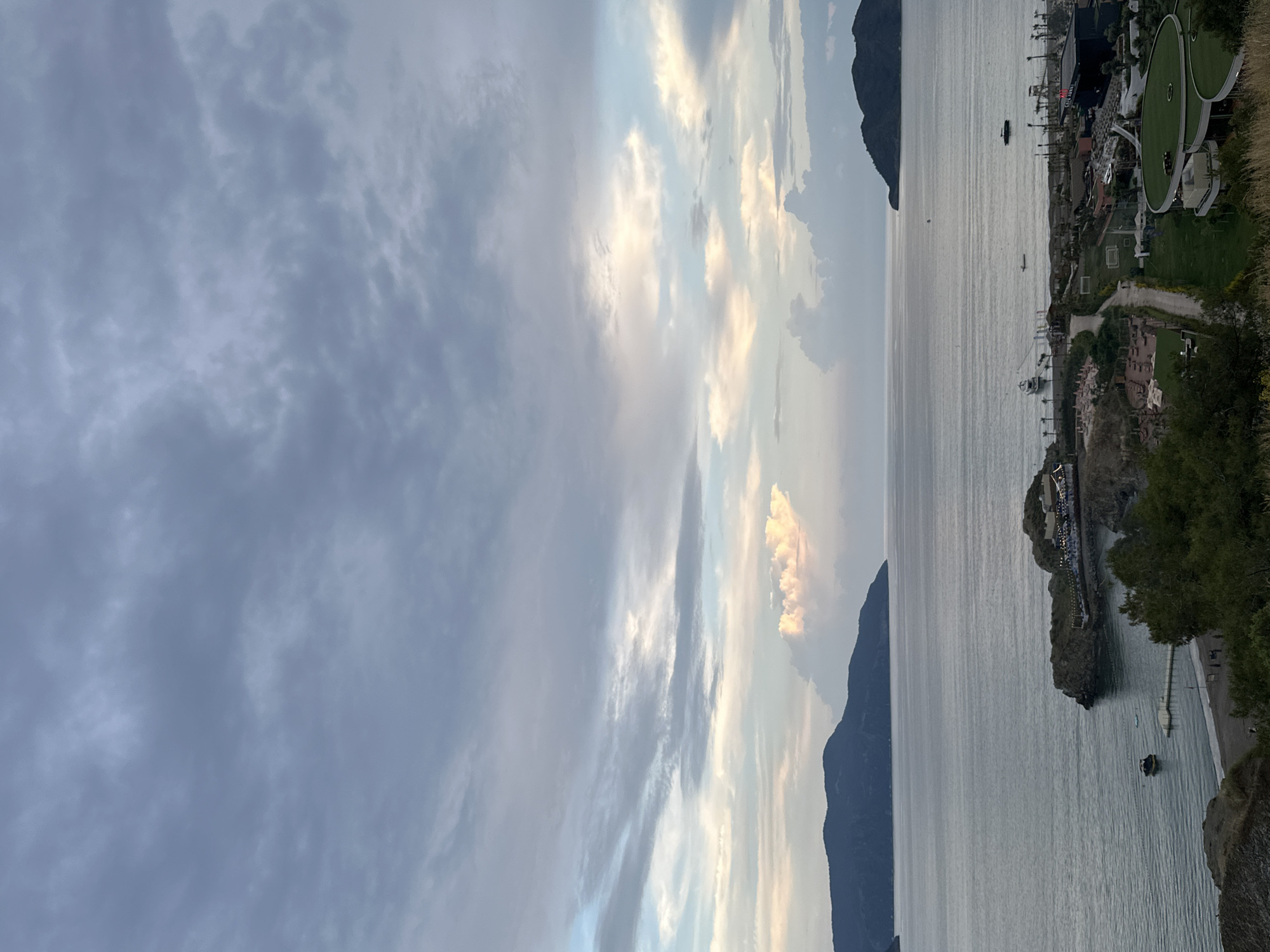 The image depicts a coastal scene in Fethiye, Turkey, as viewed from an elevated position, likely a cliff or hillside. The sky is predominantly cloudy, with varying shades of gray and hints of blue. The water, presumably the Mediterranean Sea, extends towards the horizon and is dotted with a few boats. On the right side of the image, a portion of the shoreline is visible, featuring vegetation, buildings, and some urban development. The land appears rocky and rugged in some areas, while other parts show signs of landscaping and manicured gardens. Specific details about people or actions are not discernible.