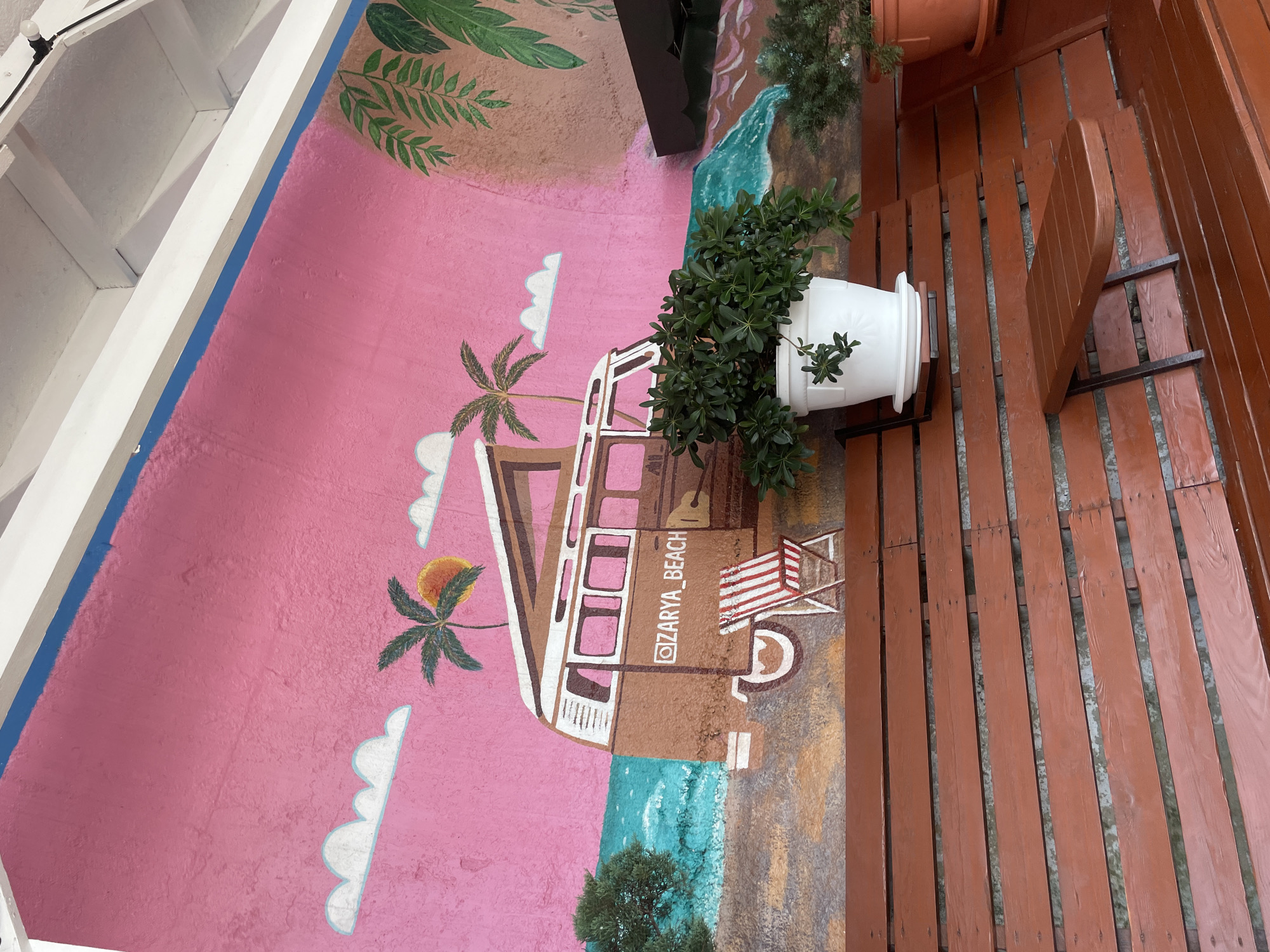 The image depicts a vibrant mural on a wall, rendered in a distinctly retro and whimsical style. The mural's backdrop is a bright pink sky, adorned with stylized white clouds and two palm trees. A vintage camper van, painted in earthy tones of brown and white, is a central element, positioned as if parked on a beach. The van features the Instagram handle "@ZARYA_BEACH" painted on its side. In front of the van, a classic red and white striped deck chair sits on a sandy, painted ground, hinting at a beachside setting. The sea, depicted with energetic turquoise waves, is also part of the mural.
In the foreground, a rustic wooden bench with a reddish-brown finish runs horizontally across the frame, suggesting an outdoor seating area. A small, brown wooden tray table is attached to the bench. A large white planter containing a lush green plant is mounted on the wall above the bench, and to its right, another orange planter with a smaller green plant is visible. The overall scene evokes a relaxed, summery atmosphere, likely a decorative element of a cafe, bar, or public space. The mural's style, with its bold colors and simplified forms, suggests a playful and inviting ambiance.
While the image does not explicitly state its location, a sign on the mural mentions "@ZARYA_BEACH," which is a reference to a beach in Sochi, Russia. This suggests the mural is located in or near Sochi.