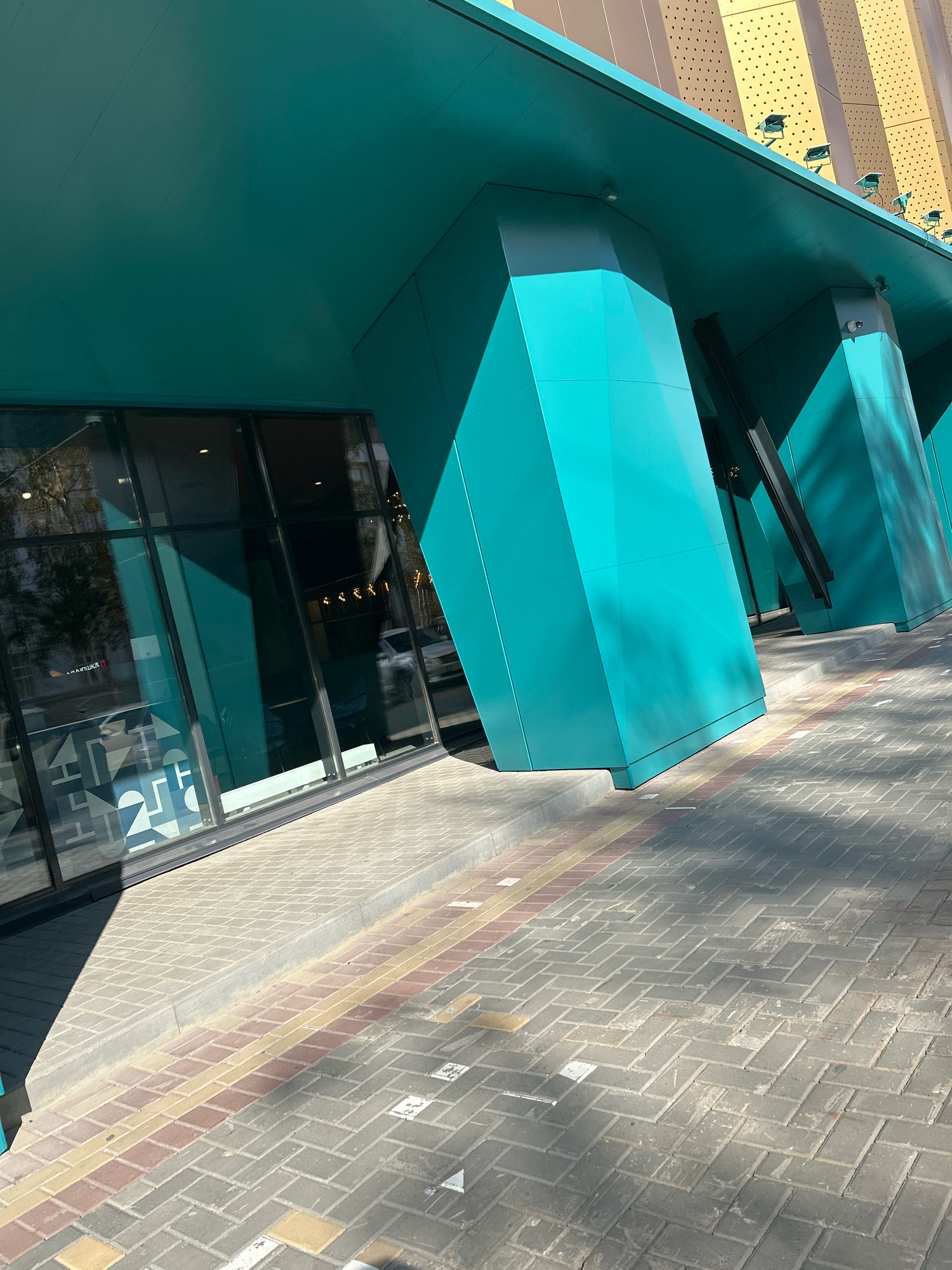 An exterior shot captures a modern building with teal-colored angled pillars and overhangs. The building features large glass windows with reflections of trees and possibly a car. The foreground shows a sidewalk paved with interlocking grey, red, and yellow bricks, indicating a pedestrian walkway. Shadows from the building stretch across the sidewalk, suggesting a sunny day. The architectural style suggests a commercial or public space. The building's facade is partly adorned with gold-colored panels featuring a pattern of small perforations, with several exterior lights mounted along the top edge. The overall scene conveys a contemporary urban environment. This specific location is the Tomsk State University of Architecture and Civil Engineering in Tomsk, Russia.