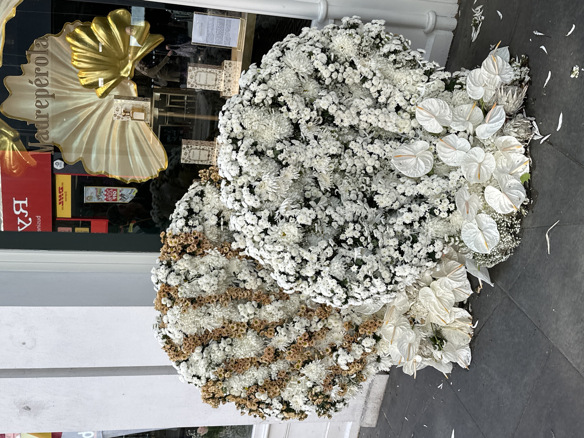 Two floral arrangements are positioned on the ground next to a storefront. The arrangement closest to the storefront is largely composed of white flowers, including what appear to be chrysanthemums, baby's breath, and white anthuriums. The second arrangement is also composed of white flowers, with sections of brown flowers interspersed. The storefront behind the arrangements has a reflective glass window, revealing the interior. The window features a large golden seashell-shaped decoration. In the reflection, passersby can be seen, as well as red and yellow DHL signs and a sign for "Madreperola". The ground is a light grey color, and a small amount of floral debris litters the area. Based on the "Madreperola" sign and the presence of the DHL delivery service, the location is likely in London, United Kingdom.