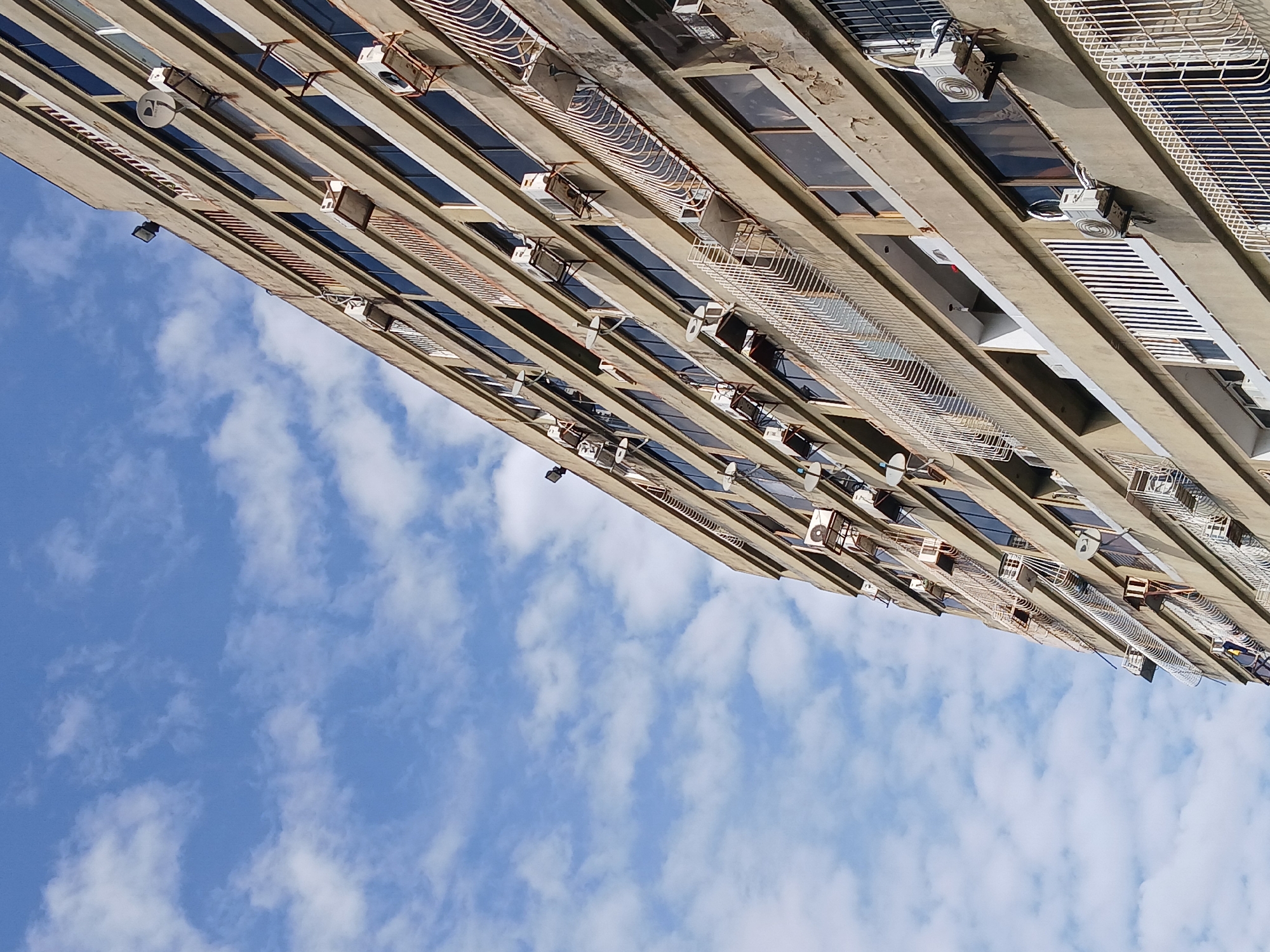 An upward-tilted view features a high-rise building in Ciudad Guayana, Venezuela against a backdrop of a partly cloudy, blue sky. The building's facade is comprised of numerous windows and balconies, most of which have window-mounted air conditioning units. The building is primarily gray concrete. The photo is taken at a low angle, giving the building a towering perspective.