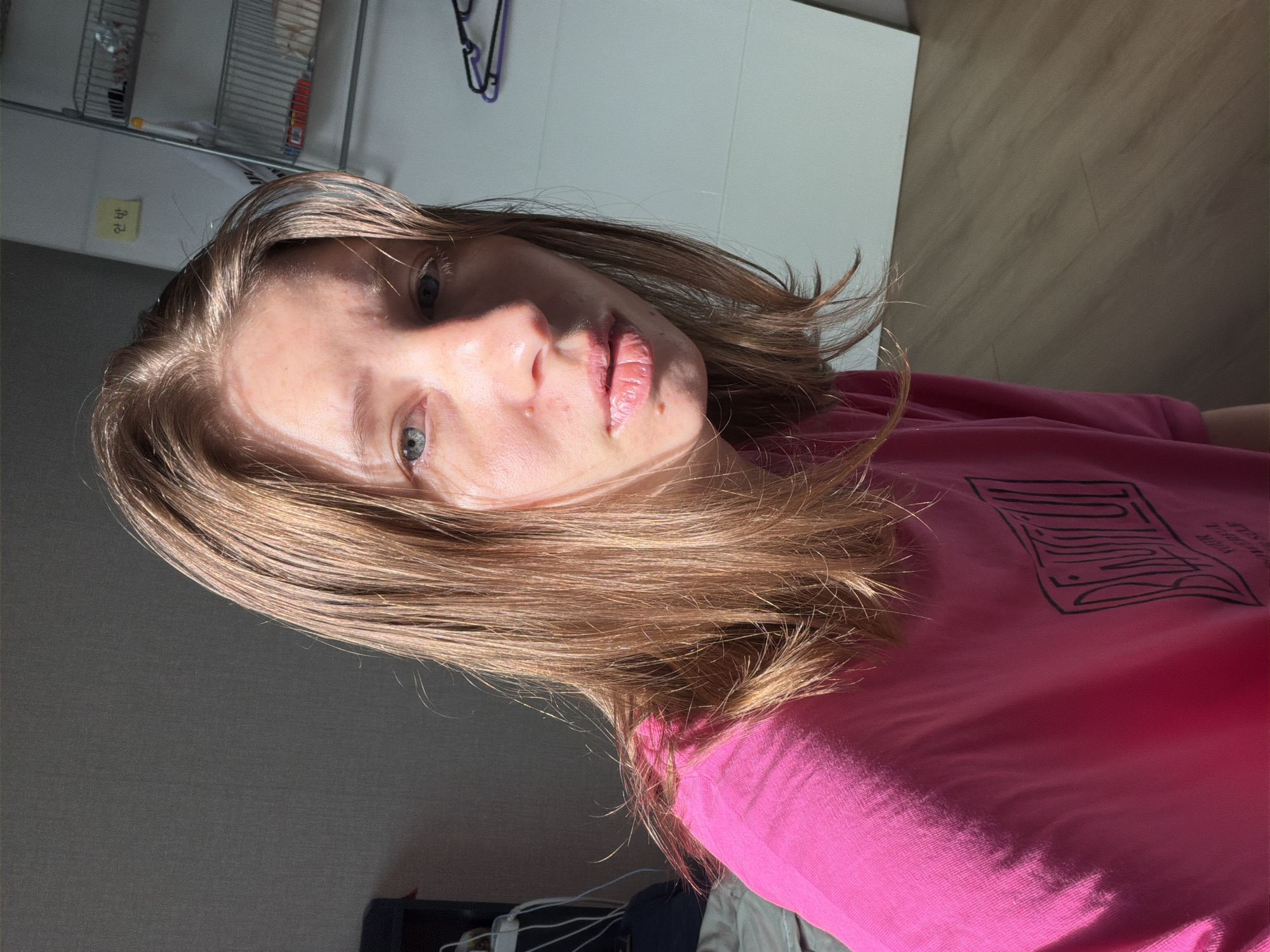 The image is a medium shot of a young Caucasian woman with shoulder-length blonde hair, looking directly at the camera. She is wearing a bright pink t-shirt with black text on the back. The woman's face is partially illuminated by natural light from the right, casting shadows on the left side of her face. Her expression is neutral. In the background, to the right, is a white closet door and a silver metal shelf unit with various items on it. A yellow sticky note with Korean text is affixed to the wall above the shelf. To the left of the woman, a dark object, possibly a piece of furniture, and some white cords are visible. The floor is made of wood planks. The overall environment appears to be an indoor residential setting. The image was taken in Dongducheon-si, South Korea.