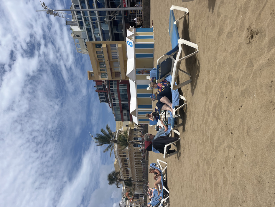 The image shows a sandy beach with people relaxing on lounge chairs. The scene is set in Las Palmas de Gran Canaria, Spain, at the coordinates 28.143, -15.433. In the background, there are buildings with balconies, palm trees, and a blue sky with white clouds. Three people are sitting on lounge chairs and reading. The scene is peaceful and relaxing, with the beachgoers enjoying the sunny day.