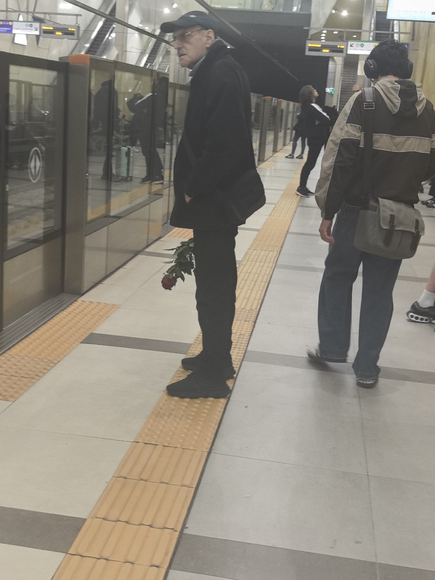 The image shows a man in a black coat and cap standing on a tactile paving surface at a metro station. He is holding a red rose. Other people are visible in the background, waiting on the platform. The station has platform screen doors.