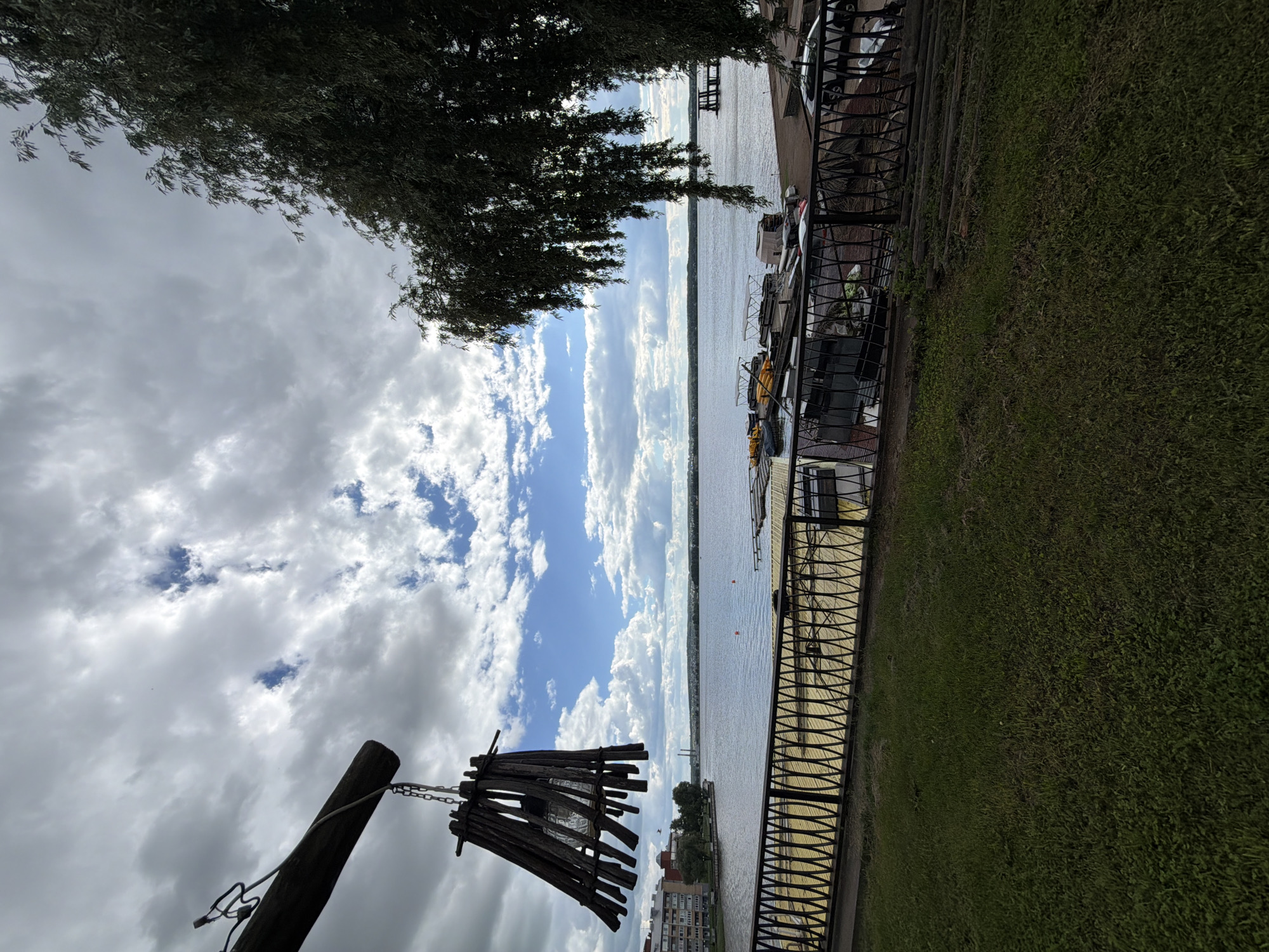 The image depicts a waterfront scene in Votkinsk, Russia, likely during the daytime under a cloudy sky. In the foreground, a grassy slope leads down to a black ornamental metal fence. Beyond the fence is a paved area with yellow markings, possibly a promenade or walkway. Across this area, a body of water, likely a river or lake, stretches out. Several boats are docked or moored along a pier on the water. In the distance, across the water, the Votkinsk dam and hydroelectric power station are visible, characterized by large concrete structures and smokestacks. The architecture of buildings on the far bank suggests an urban or semi-urban setting. The sky is filled with a mix of grey and white clouds, with patches of blue sky visible. A rustic wooden fixture, possibly a lamp or decorative element, hangs from a wooden beam in the upper left corner of the frame, with a clear glass bulb visible within its wooden structure. A large, leafy tree with drooping branches partially obscures the view on the right side of the image. The overall impression is one of a scenic, slightly overcast day at a riverside location.