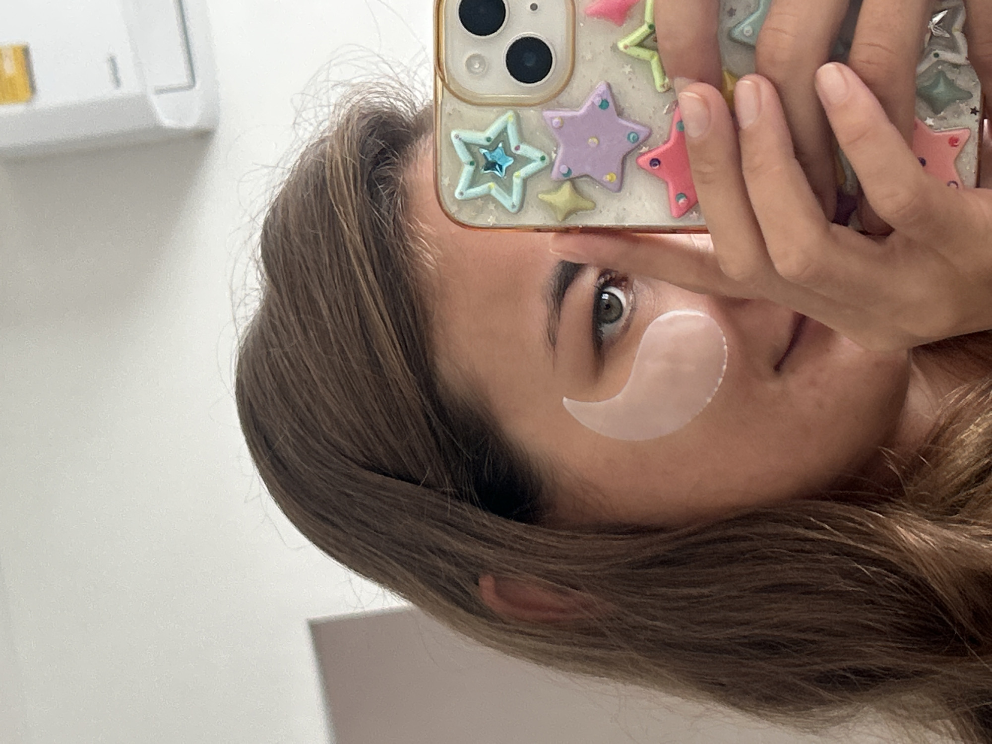 A young woman with brown hair is taking a selfie. She is wearing a light pink under-eye patch on her right eye. Her left eye is visible, and she is looking at the camera. She is holding a smartphone with a clear case decorated with colorful star stickers. The background appears to be an indoor setting with a white wall and a light-colored vent or air conditioning unit on the upper right. The image is cropped to focus on the woman's face, hair, and the phone. No specific location in Kathu, Thailand can be identified from the image.