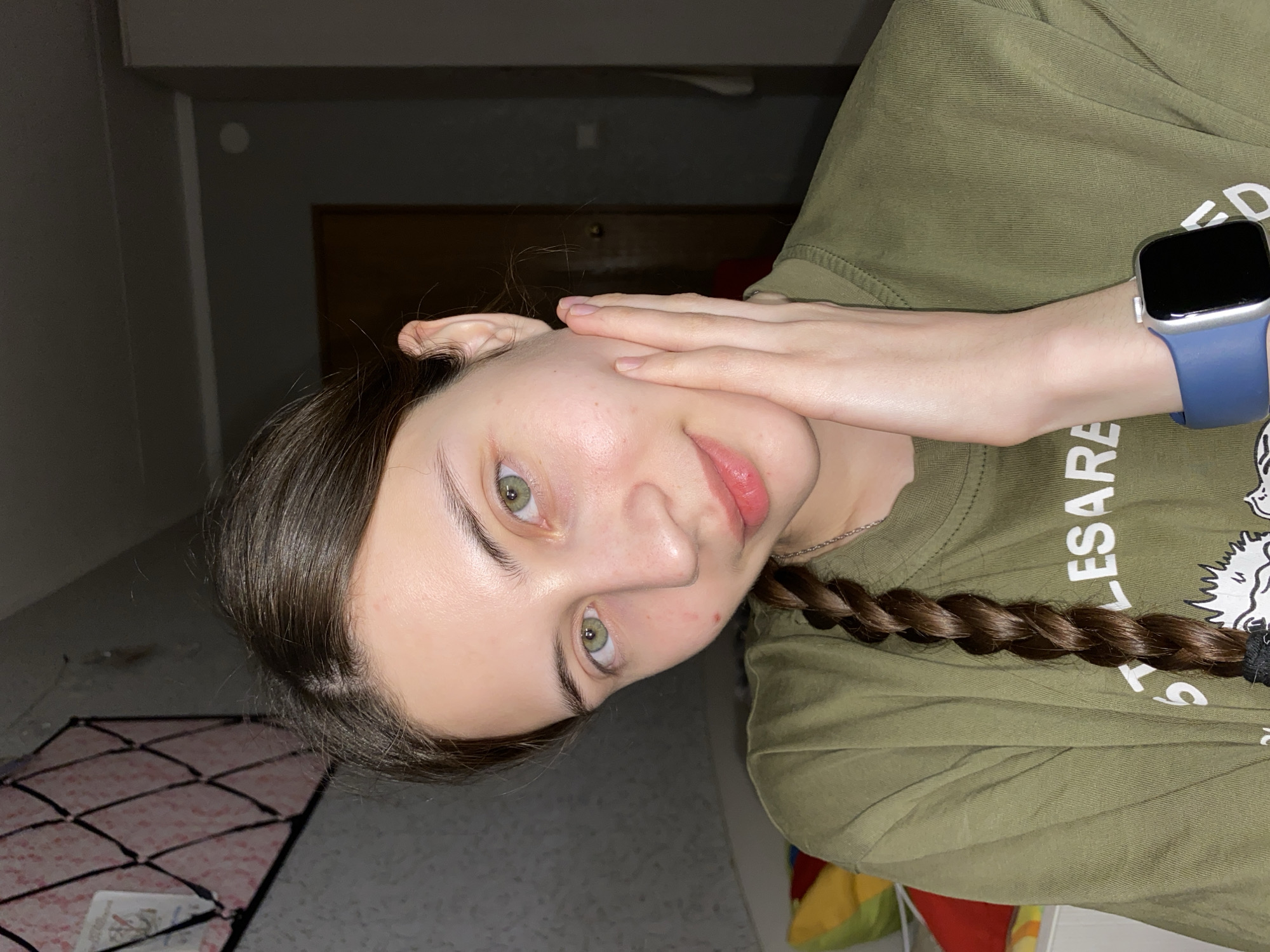 A young woman with fair skin and green eyes is oriented at an angle. She has brown hair styled in a braid. Her hand is touching her face. She is wearing a green shirt and a blue smartwatch. The background appears to be an interior, possibly a room in a house, but details are limited. There is no information available in the image to establish its location as Votkinsk, Russia.