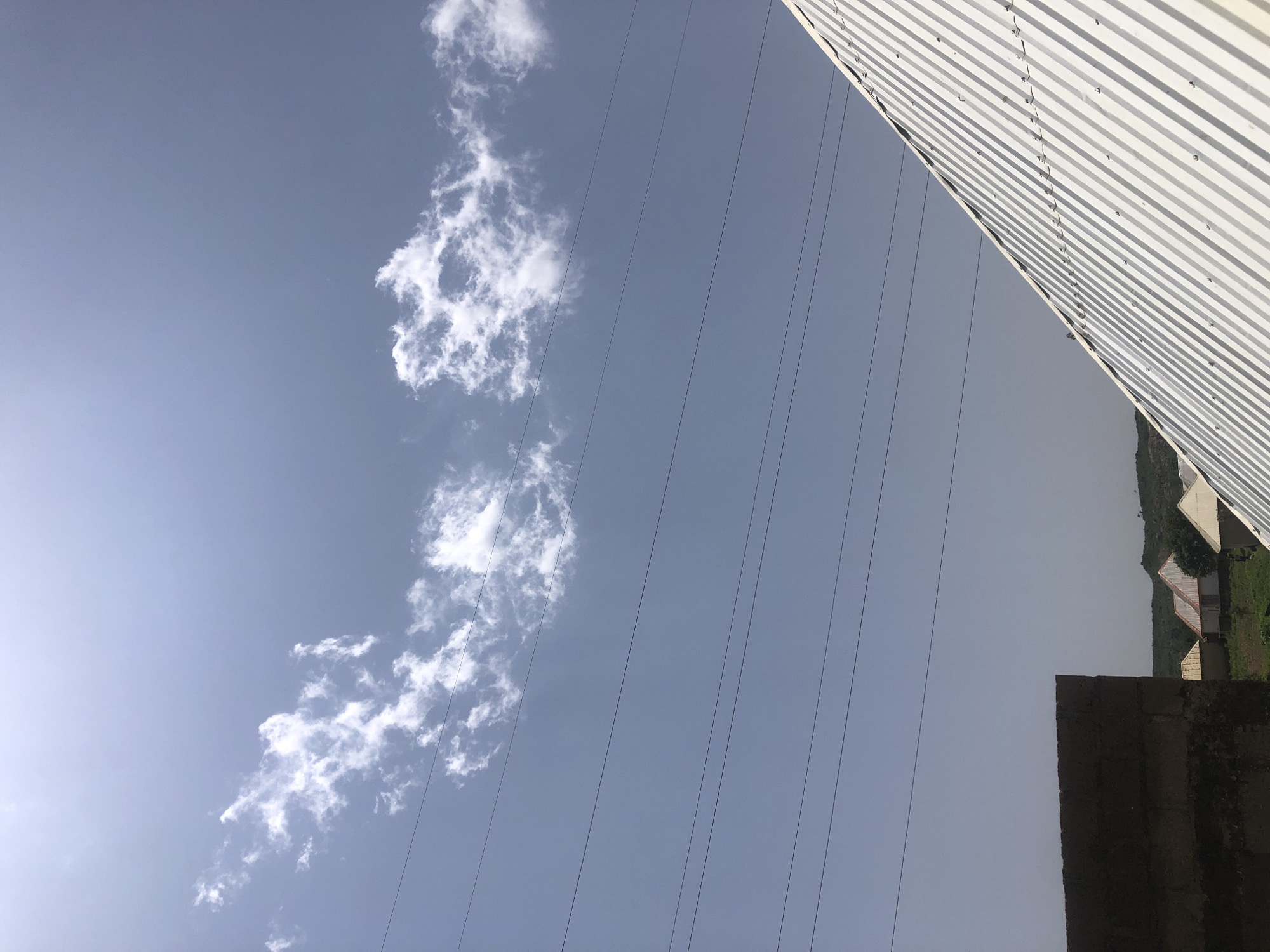 An outdoor, medium-shot, eye-level photograph captures a bright, hazy sky with scattered white clouds. Several horizontal power lines traverse the sky, dividing it into bands. In the foreground, the top of a corrugated metal roof slopes diagonally from the bottom right to the center of the frame. To the left, the top of a dark, rough-textured wall extends from the bottom left corner upwards and to the right. In the distance, a line of small, rustic buildings with peaked roofs and a faint green landscape are visible beneath the power lines. The image appears to be taken on a clear day, with the sun casting a bright light.