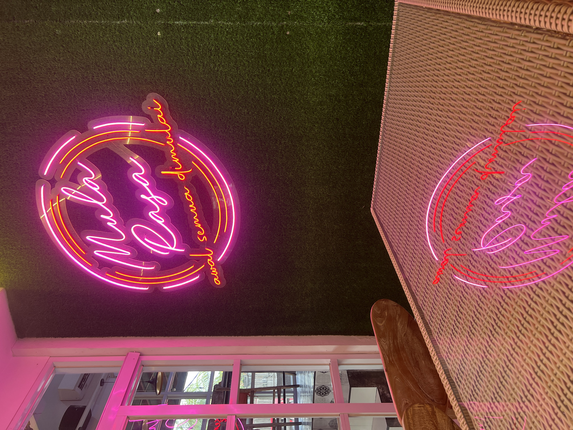 The image captures an interior scene in Sukawati, Indonesia, where a neon sign reading "Mula Rasa awal semua dimulai" is affixed to a ceiling covered with artificial grass. The sign consists of pink and yellow neon lights. The background includes a pink-framed window with visible outdoor scenery of trees and a building exterior. To the right, a section of a wicker table is visible, also bearing a neon sign of a similar design.