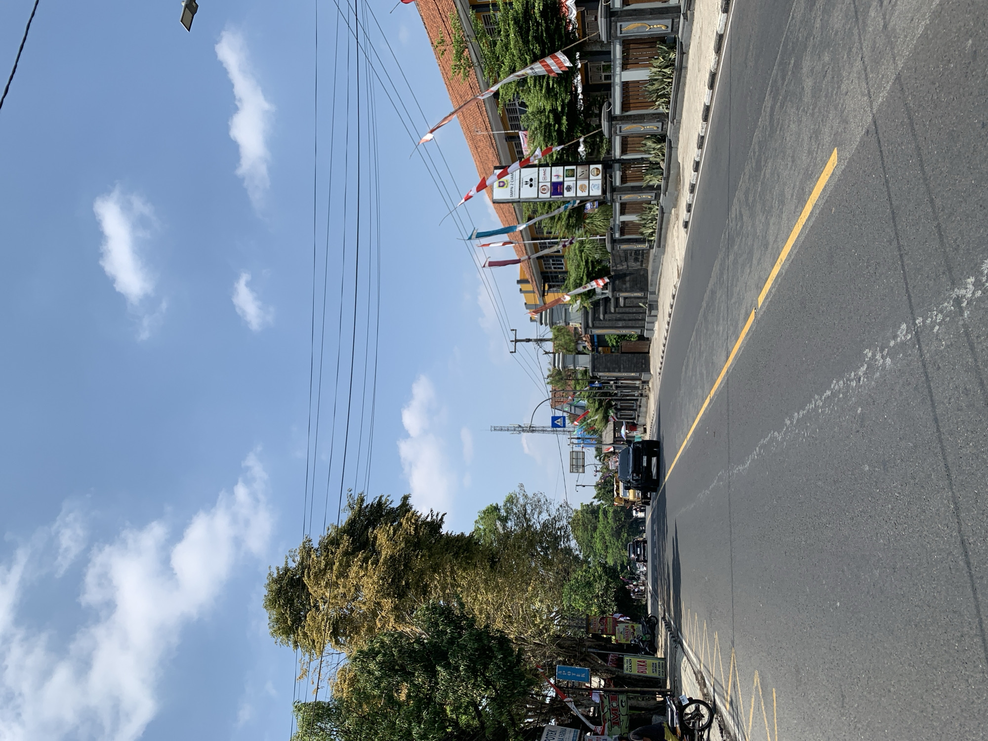 The image captures a daytime street scene in Ciamis, Indonesia. The perspective is from the middle of a two-lane asphalt road with a yellow dashed line marking the center. On the left side of the road, there's a sidewalk lined with trees, some with green foliage and one with golden-brown leaves, and various shops and small buildings with signs, including one with the text "APOTEK" and "BUKA". A parked motorcycle is visible on the sidewalk. On the right side of the road, a sidewalk is bordered by a decorative fence and a building with an orange roof, adorned with Indonesian flags. Several other flags and banners are also visible along the street. Traffic includes a black SUV, a yellow truck, and other vehicles further down the road. A cellular tower is visible in the distance, along with a pedestrian crossing sign. The sky is blue with scattered white clouds.