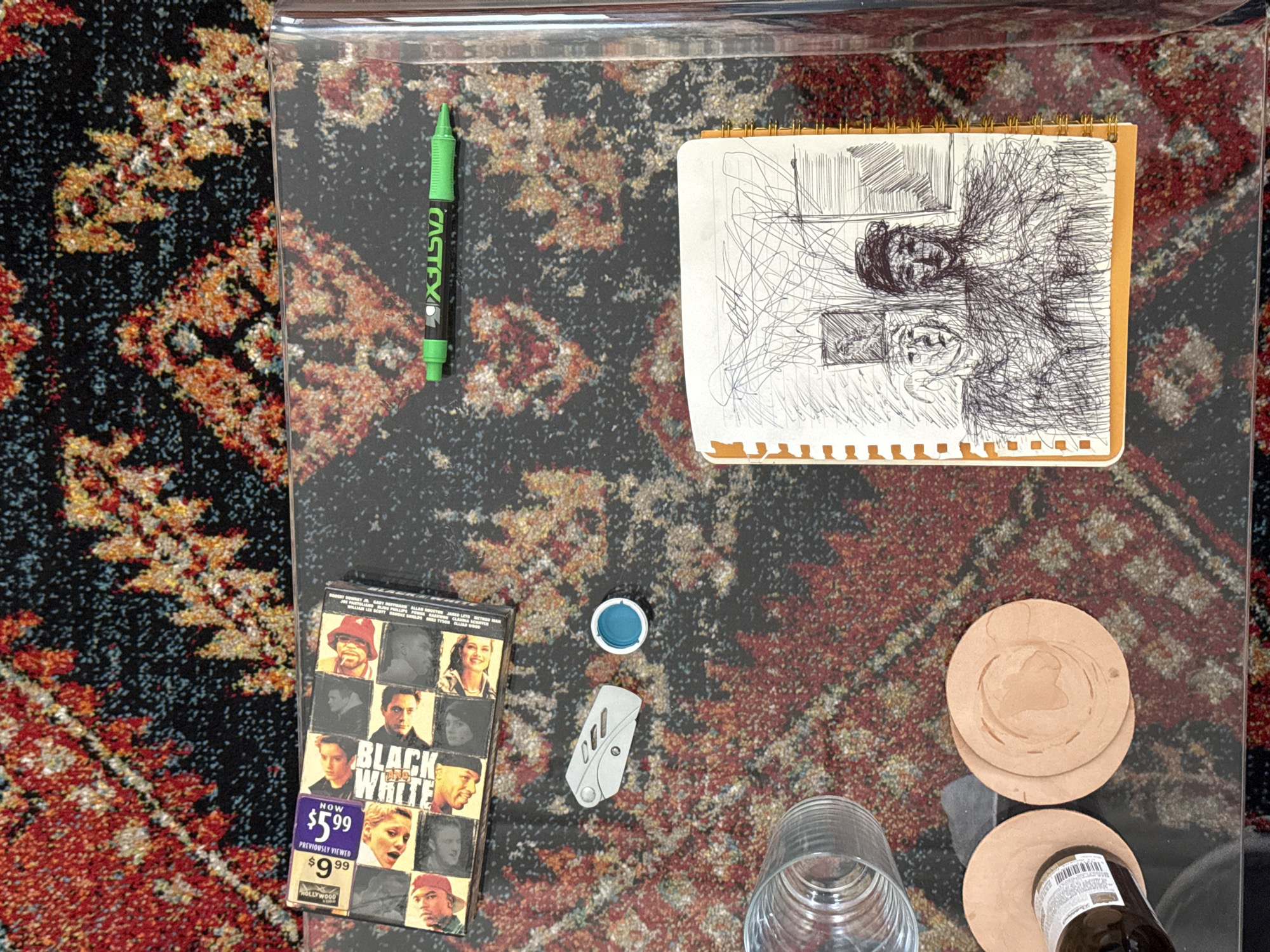 On a glass coffee table, amidst a patterned rug, is an assortment of items: a green Castelli pen, a sketchpad with a portrait drawing, a VHS copy of the film "Black and White", a small blue cap, a pocket knife, a drinking glass, and a stack of leather coasters supporting a partially visible beer bottle. There is no explicit evidence to suggest that this is in New York City.