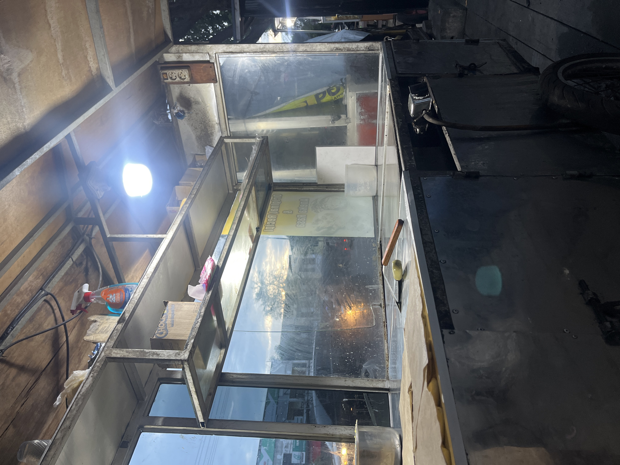 This is a low-angle, close-up, eye-level medium shot of a food cart at dusk in Banda Aceh, Indonesia. The food cart is made of stainless steel and wood, with a glass window that reflects the sky and the surrounding environment. A bright light hangs from the ceiling of the cart, illuminating the interior. On the shelves, there are boxes of food and a bottle of cleaning spray. A metal pot is visible on the left side of the cart, and a piece of parchment paper is spread out on the counter. The cart is positioned on a wooden walkway, and the background shows a street scene with trees and buildings. The overall impression is one of a bustling street food stall in a tropical city.