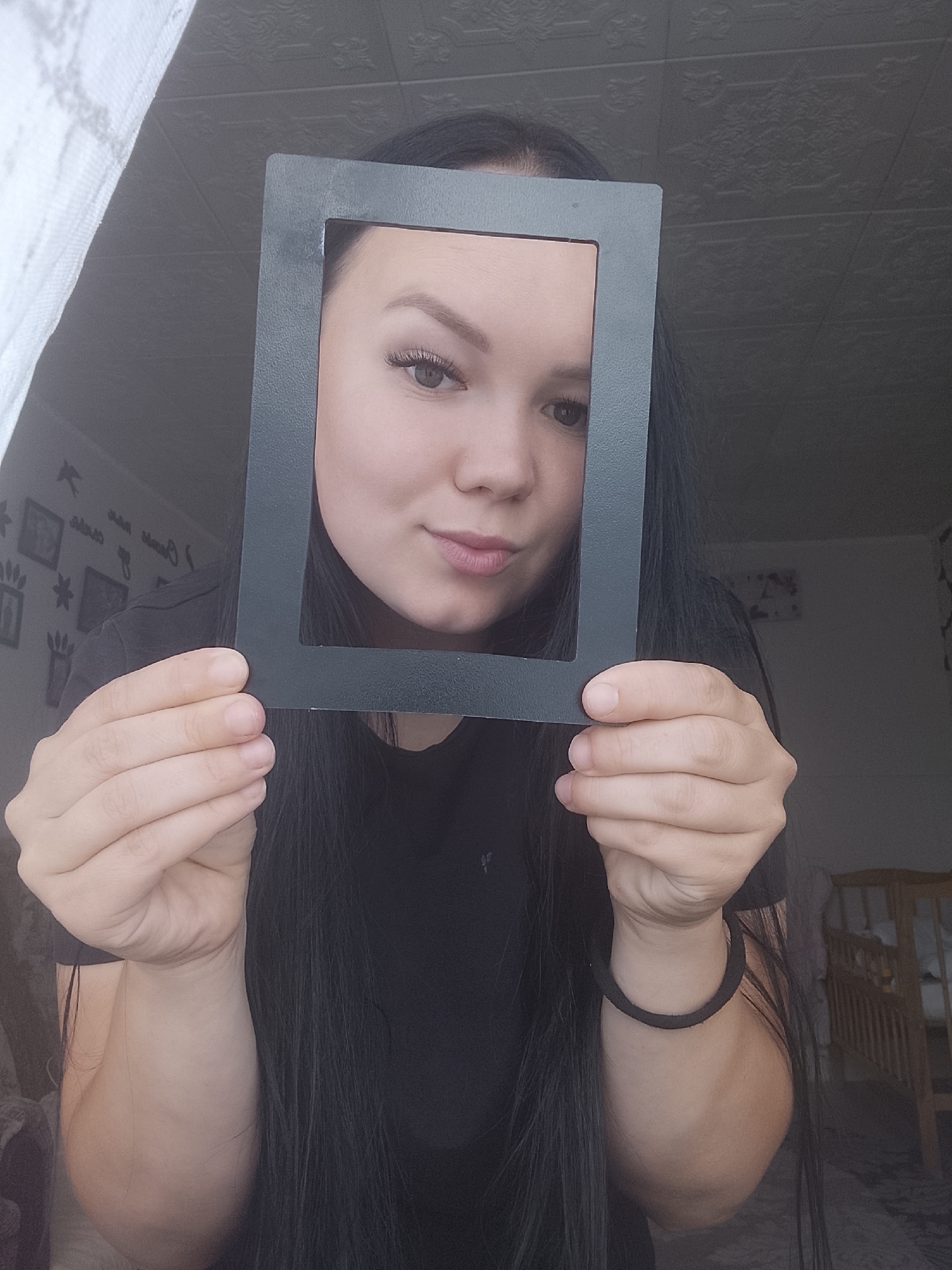 An individual with long, dark, straight hair, wearing a dark top, is centered in the frame, holding a matte black rectangular frame with both hands at eye level. Her face is framed by the object, displaying light-colored eyes (likely hazel or light brown), well-defined eyebrows, and enhanced eyelashes. Her lips are slightly parted, forming a subtle smirk or smile, and she is looking directly at the viewer. A black hair tie is visible on her right wrist.
The background depicts an indoor domestic setting. To the upper left, a white, textured fabric is partially visible. Below it, a light-colored wall with various dark silhouette decals (birds, text, abstract shapes) is present. The upper background features a ceiling with embossed or textured tiles. To the right, the wall is darker, and a light-colored wooden structure, resembling a crib or bed frame, is partially visible in the lower right corner. The lighting is dim and ambient. The specific geographical location, such as Kuyeda, Russia, is not discernible from the visual evidence.