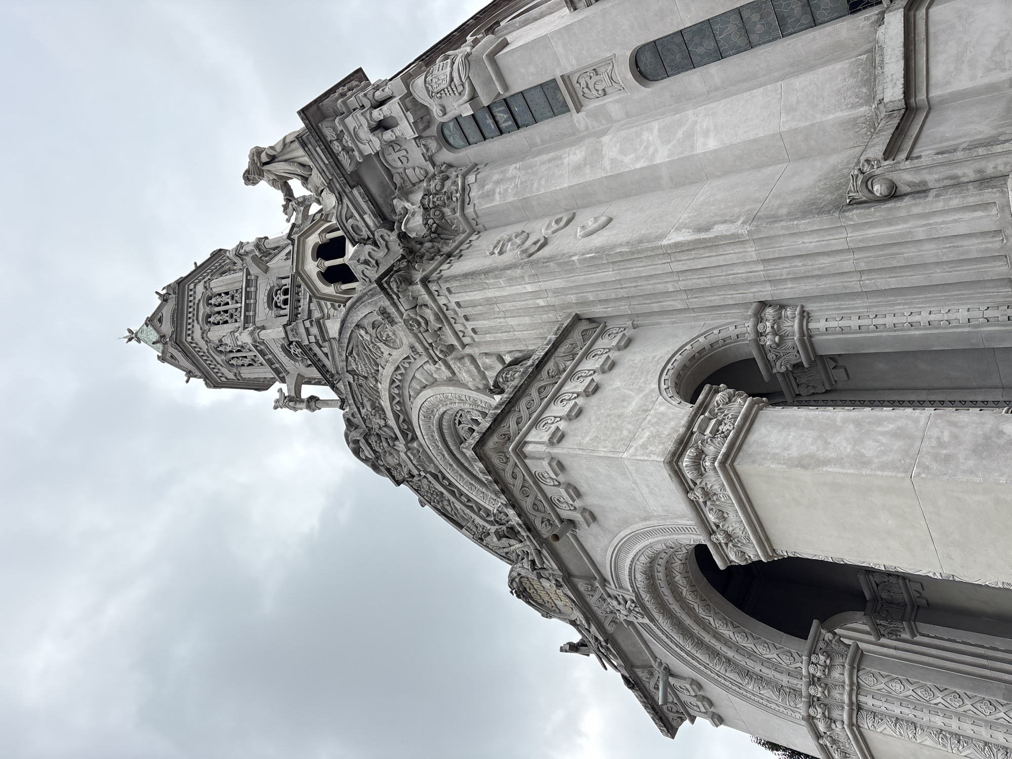 This is a low-angle, full shot of the exterior of the Mausoleum of the Counts of Próscio at the Consolação Cemetery in São Paulo, Brazil. The sky above is overcast and cloudy, with a muted gray hue. The mausoleum is constructed of a light-colored stone, possibly marble or granite, and features intricate architectural details.
The left side of the image shows a large arched entryway with decorative carvings around the arch and on the supporting columns. The columns are fluted and topped with ornate capitals. Above the arch, there is a frieze with elaborate scrollwork and a circular relief. To the right of the entrance, the mausoleum rises dramatically. A prominent tower with multiple levels and windows is visible, topped by a pointed spire. Statues are present on ledges and architectural features of the mausoleum. One statue, appearing to be a robed figure, stands prominently on a ledge near the tower, holding a smaller figure. Other decorative elements, such as gargoyles or decorative carvings resembling faces, are present on the building's facade. Stained glass windows with geometric patterns are visible on the right side of the structure. The overall impression is one of grandeur and historical significance.