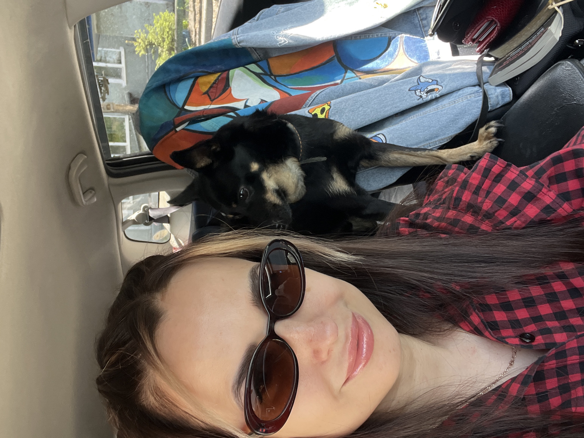 A young woman takes a selfie from inside a car, holding the camera overhead. She is positioned on the left side of the frame, smiling slightly with glossy lips, and wearing dark sunglasses and a red and black plaid shirt. Her long, dark brown hair, with lighter highlights, falls around her shoulders. A thin gold-colored necklace is visible around her neck.
Behind her, a black and tan dog lies calmly on what appears to be a light blue denim jacket. The jacket features colorful, abstract designs and cartoon shark logos; one logo clearly shows a shark eating a pizza slice with the word "PIZZA" visible above it, and another shark is depicted wearing a cap.
The interior of the car is visible, including a beige ceiling or pillar on the left, a grab handle, and part of a car seat headrest. Behind the dog and jacket, a red wallet, a book, and other items are clustered. Through the car window or sunroof, sunlight illuminates the scene, and outside, some green foliage and parts of a building or structure with a ladder can be seen, indicating it is daytime. The dog appears relaxed, and the overall mood is casual and comfortable.