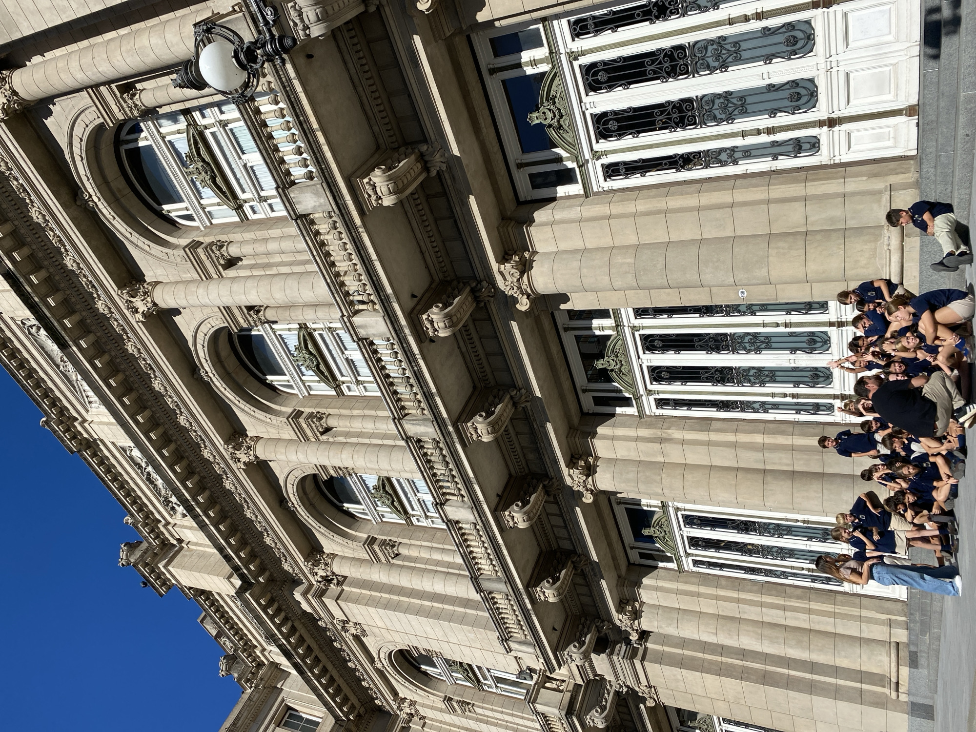 This low-angle photograph captures the ornate stone facade of a grand building under a clear blue sky, set in Buenos Aires, Argentina. The building displays classical architectural elements, including multiple stories with decorative pilasters, columns, intricate cornices, and detailed friezes. Arched windows on the upper levels and rectangular windows on the lower floors feature prominent white frames and decorative black wrought-iron grilles. An antique-style street lamp with a white globe is affixed to the building's exterior.
In the foreground, a group of approximately 15-20 young individuals, most of whom are dressed in dark blue polo shirts, are gathered on the steps or sidewalk directly in front of the building. They appear to be an organized group, possibly students on an outing. Many are crouched or sitting close together, looking towards the right side of the frame, suggesting they are posing for a photograph or listening to a guide. An adult man in a dark shirt and khaki pants is seen crouching with the group, while another adult woman in a light top and blue jeans stands slightly apart to their left.
The bright sunlight casts distinct shadows across the facade, indicating a clear, sunny day. The overall atmosphere suggests a daytime visit or tour. No legible text is visible on the building or among the people.