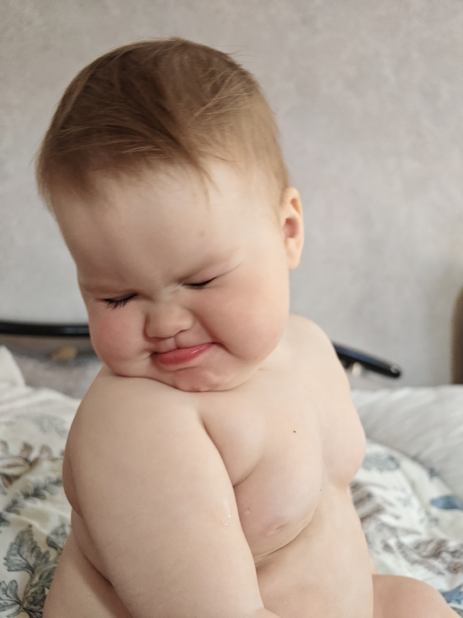 A close-up shot captures a baby with fair skin and light brown hair, who appears to be grimacing or scowling. The baby's eyes are closed in a squint, and the mouth is turned down in a pout. The baby is mostly undressed, exposing shoulders and part of the torso. The baby is sitting on a bed, as indicated by the patterned bedding in the background. The location is Chaikovsky, Russia.