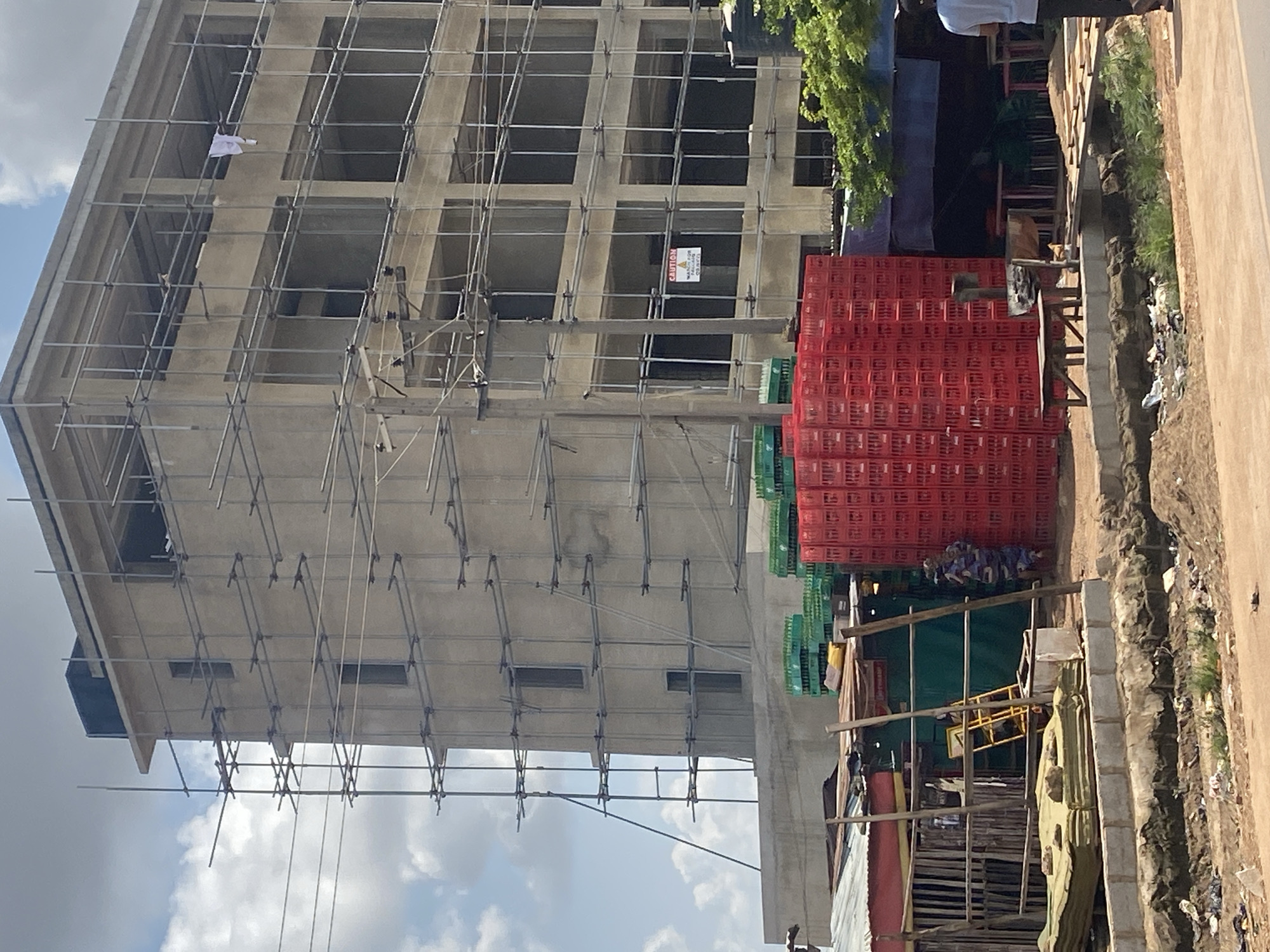 An unfinished concrete building, encased in scaffolding, dominates the frame in Ado, Nigeria. The structure is multi-storied, with open window frames suggesting ongoing construction. Below the building, stacks of red and green plastic crates are piled high. In front of the crates, a small, makeshift stall with a corrugated metal roof is visible, featuring a yellow ladder and some bundled materials. A person in a blue and patterned outfit stands partially obscured by a wooden post near the stall. The foreground shows a dirt road with a trench running alongside it, containing stagnant water and debris. A mature tree with green foliage is seen on the right side of the image. The sky above is partly cloudy.