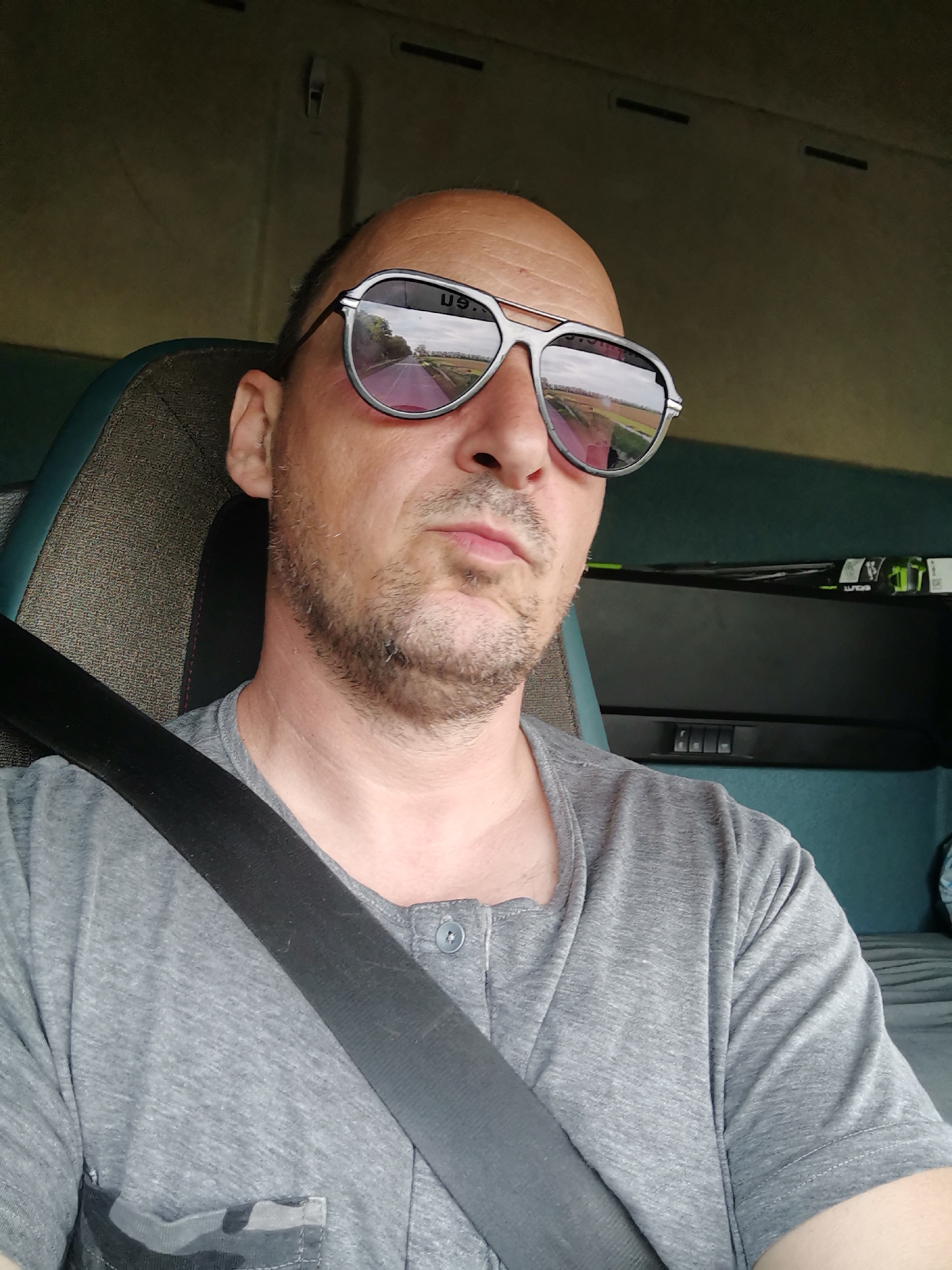 A man is sitting in a vehicle wearing a seatbelt and sunglasses that reflect a rural road and fields. He has a shaved head and stubble, and is wearing a gray t-shirt with a pocket. The interior of the vehicle appears to be a truck cab. The location is not explicitly indicated as being in Stozher, Bulgaria in the source image.