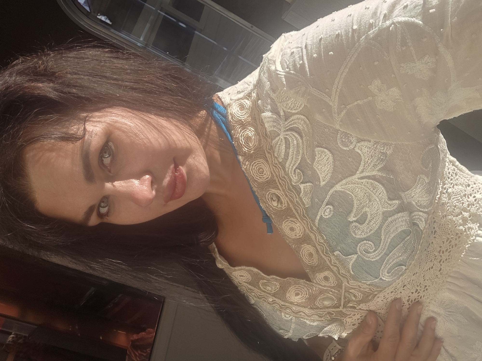 A woman is posing for a selfie in a dimly lit room, with sunlight casting shadows on her face. She has long, dark hair and is wearing a white lace dress with a blue undergarment visible. Her hand is resting on her chest. The background shows a blurred reflection in what appears to be a window or mirror, with some interior details visible. The scene is likely indoors, possibly in a residential setting or a hotel room. The exact location in Sochi, Russia is not discernible from the image.
