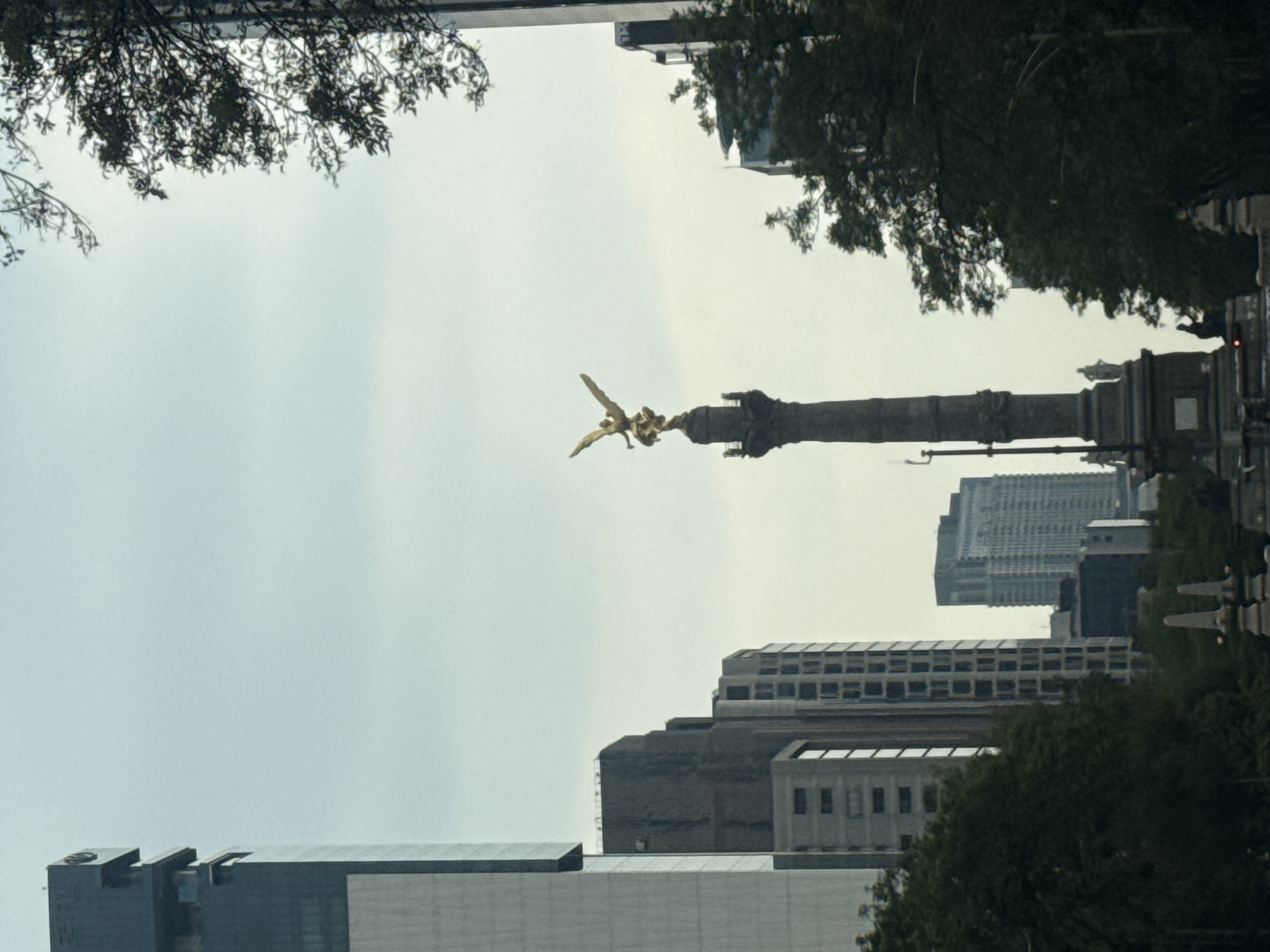 The image captures the Angel of Independence monument in Mexico City, viewed from a low angle through trees and buildings. The monument's golden angel figure is prominent against the overcast sky. Modern buildings form a backdrop. People are visible along the base of the monument. The foreground features trees.