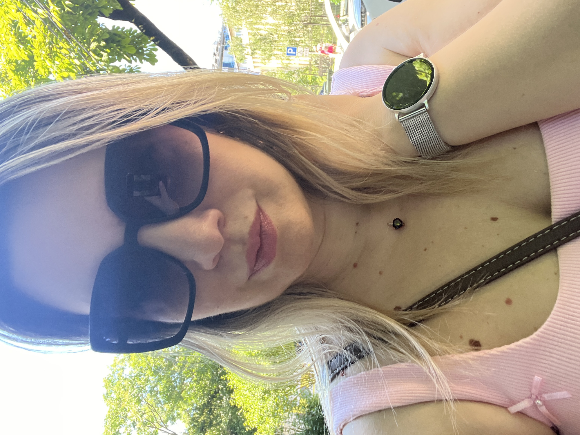 The image is a selfie of a woman taken outdoors during the daytime. She is wearing large, dark sunglasses and a pink top with a ribbon bow detail. On her wrist, she wears a silver mesh band watch with a circular face that reflects the greenery of the surroundings. Her blonde hair is visible around her face. A dark brown strap, possibly from a bag, is diagonally across her chest, and a small necklace with a dark pendant is visible at her neckline. The background shows lush green trees and foliage, suggesting a sunny, warm day. A street sign with the letter "P" and a red symbol is visible in the blurred background, hinting at a public outdoor space like a park or street in a city. The overall mood is casual and relaxed.