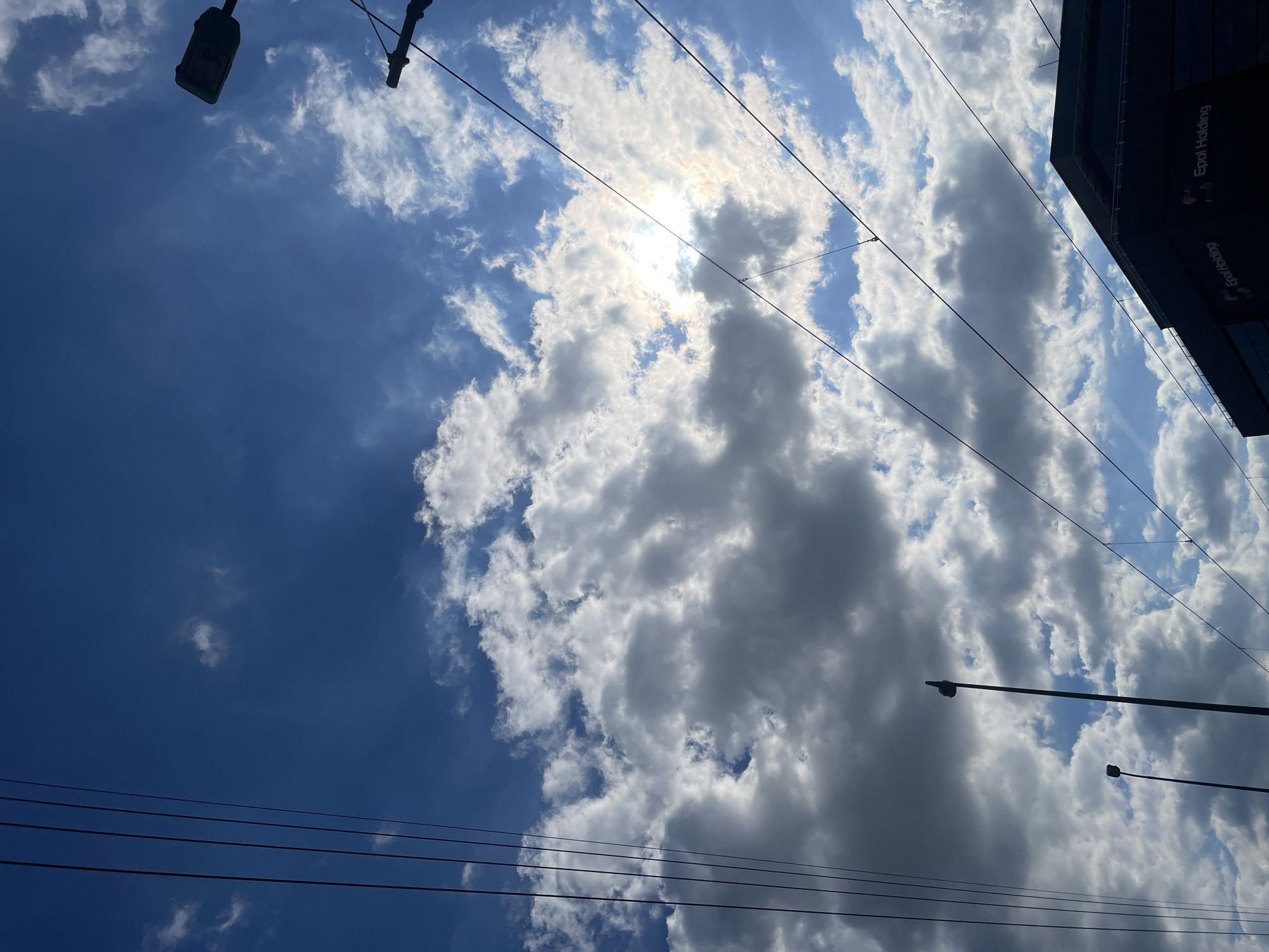 The image is an upward view of a sky filled with clouds, some dark and heavy, others lighter and whiter, obscuring the sun. Electrical wires crisscross the frame. A street lamp is visible in the upper left and another one in the lower right. In the upper right corner, a building with the words "Epot Holding" is visible. The location is Unknown, Unknown.