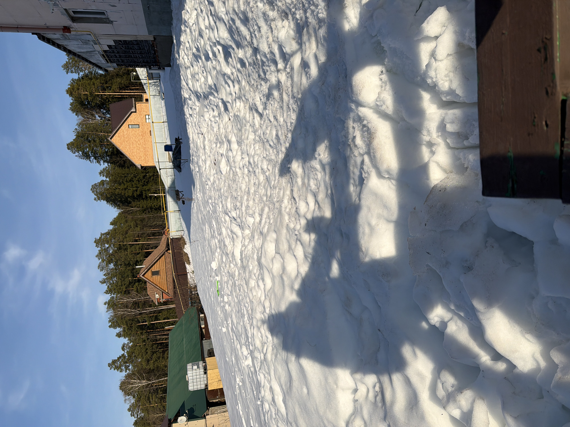 The image shows a snow-covered ground, likely in winter, in what appears to be a residential area in Chaikovsky, Russia. Several houses are visible in the background, including one with a green roof and another with an orange brick exterior. A metal fence runs along the property lines. The sky is clear and blue. The perspective of the shot is unusual, appearing to be taken from a slightly elevated and angled position. Large shadows fall across the snow-covered ground. The snow is uneven and textured. A wooden structure appears in the lower-right corner of the image.