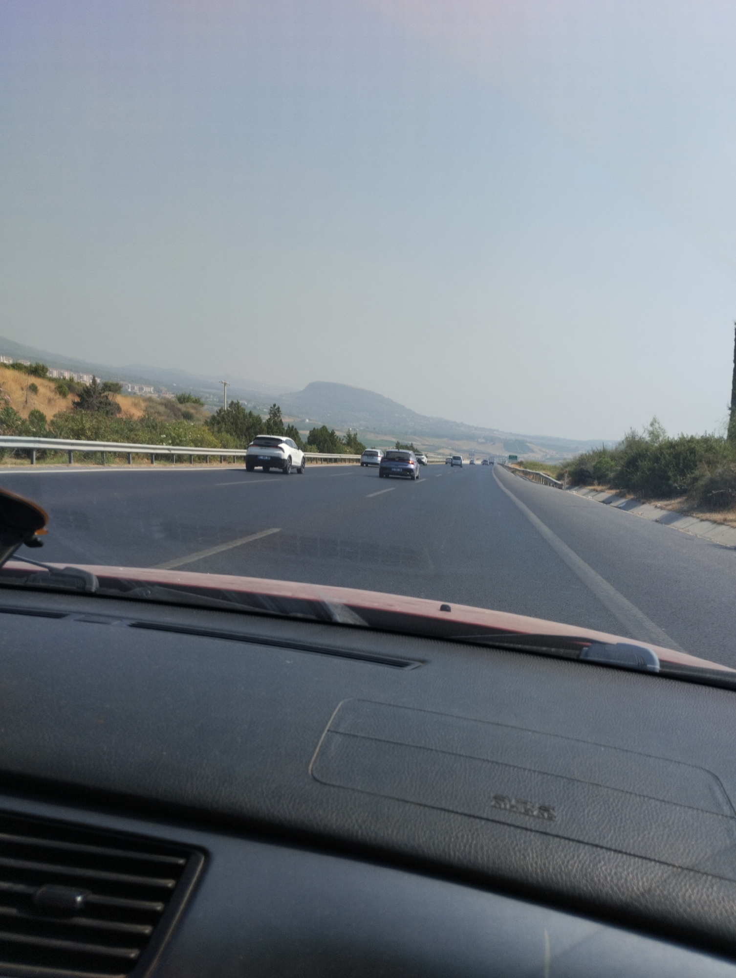 A view from inside a vehicle shows a highway with multiple cars traveling in the same direction. The road is paved and has lane markings. Guardrails line the side of the highway, with vegetation and hills in the background. The sky is clear and pale. The interior of the vehicle is visible in the foreground, including a dashboard and part of the windshield. This scene is consistent with a highway in or near Mezitli, Turkey, given the Mediterranean vegetation and hilly terrain.