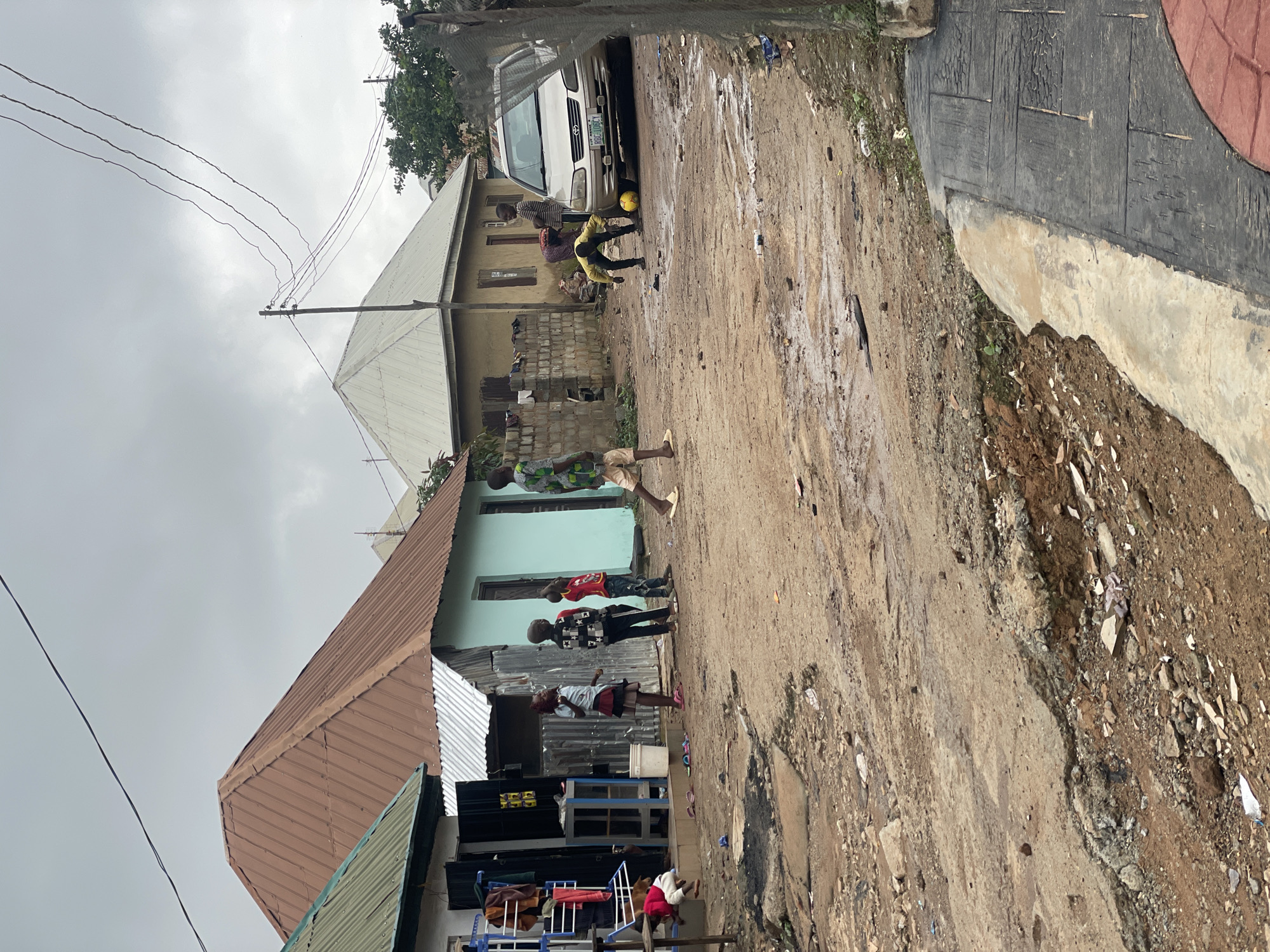 In Kurunduma, Nigeria, under a cloudy sky, several children are playing soccer in a dirt road. The road is uneven and muddy, with puddles of water in some areas. Houses with corrugated metal roofs and pastel-colored walls line the street. A silver sedan is parked on the right side of the road. One child, wearing a yellow shirt and black pants, is actively kicking the soccer ball. Other children are standing nearby, watching the game. In the foreground, a low concrete structure is visible.