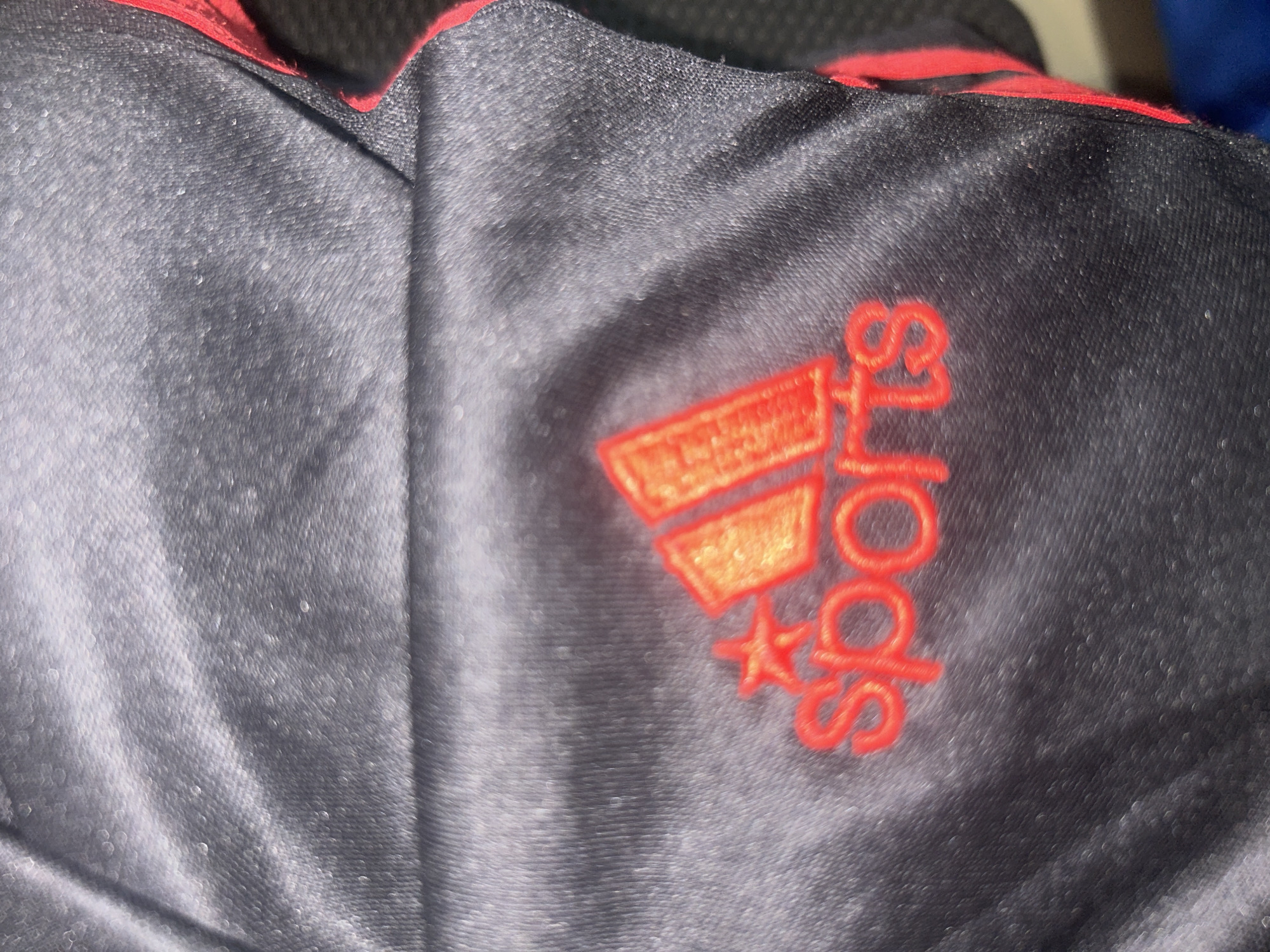 The image is a close-up shot of dark fabric with a red Adidas logo embroidered on it. The word "Sports" is also embroidered below the logo in red thread. The fabric appears to be shiny and textured, possibly sportswear. There is a red trim along the edge of the fabric. The image is taken in Ogbomosho, Nigeria. The lighting is somewhat dim, creating reflections on the fabric. There are no people or actions visible in the image.