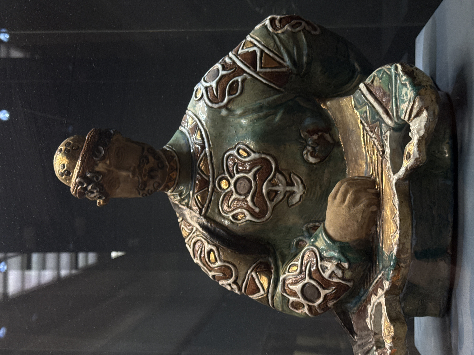 A low-angle shot shows a glazed ceramic sculpture of a seated figure playing a stringed instrument. The sculpture is displayed behind glass. The figure has a bearded face and wears a cap. His tunic is green and decorated with brown and white geometric patterns. The stringed instrument is off-white with gold accents. It's partially obscured by the figure's hand. The background is dark and out of focus, but the reflection of a building can be seen in the glass. This object appears to be an exhibit piece located in Moscow, Russia.