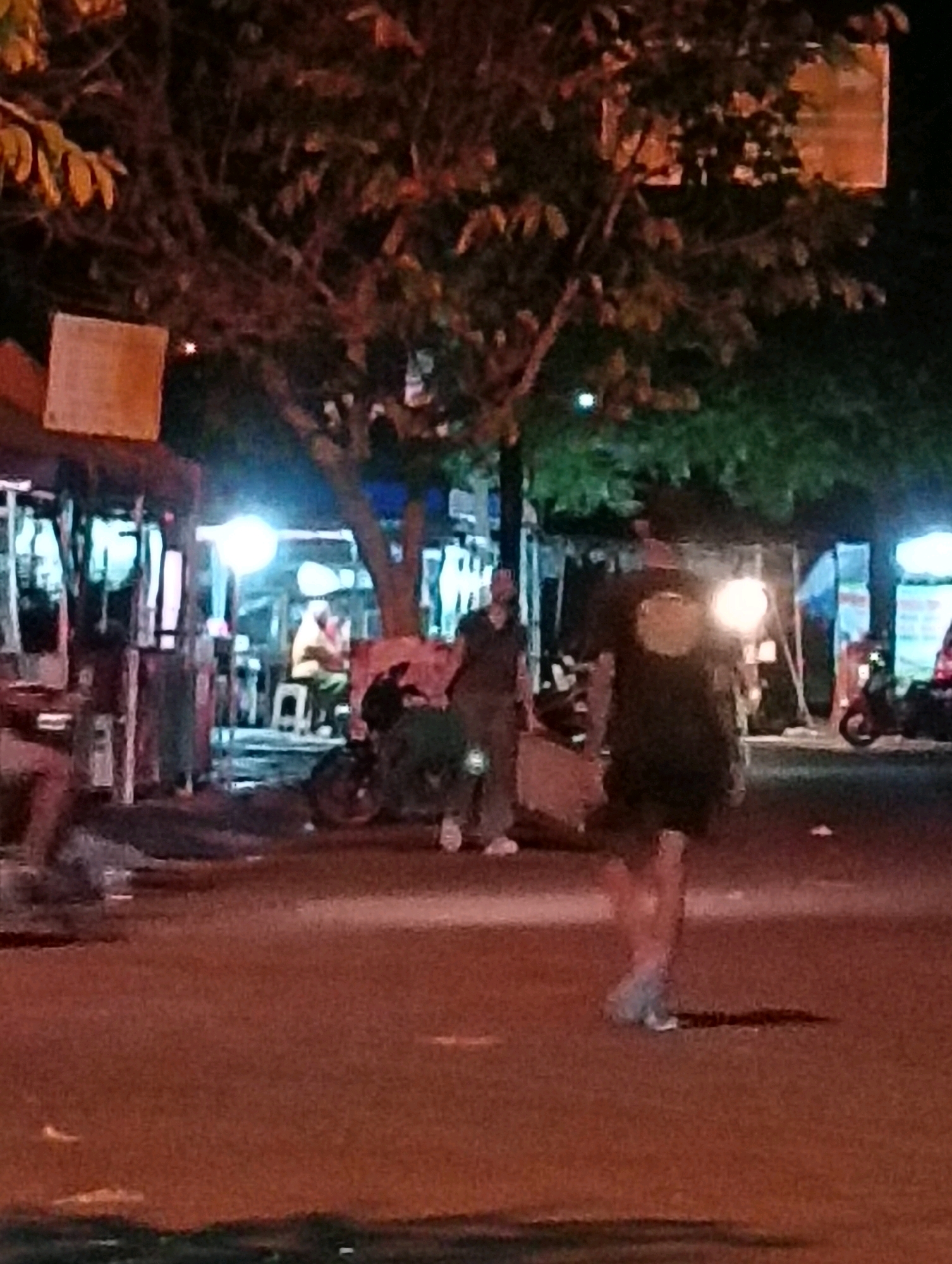 The image depicts a night scene in Purworejo, Indonesia, likely an outdoor street market or roadside stalls. In the foreground, a person is walking away from the camera, their face obscured by a bright light source behind them. They are wearing dark clothing and light-colored shoes. To the left, another person stands facing away, partially obscured by a stall. In the background, several stalls are visible, illuminated by artificial lights, with a tree overhead. A motorcycle is parked to the right of the frame, and a person is seated at a table within one of the stalls. The ground is asphalt, and the overall lighting suggests evening or nighttime.