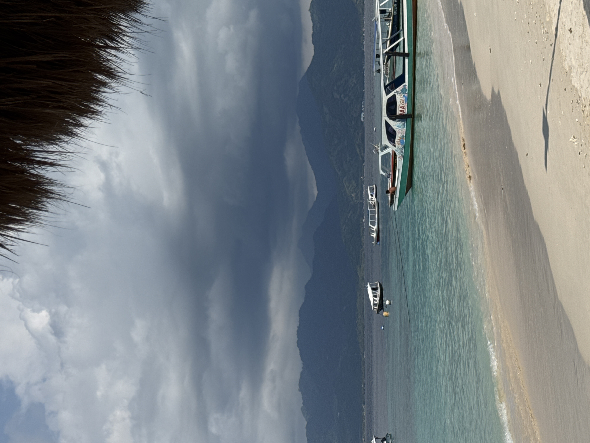 The image shows a beach scene in an unidentified location in Indonesia. A partially visible thatched roof is in the upper left corner. A sandy beach runs along the right side of the frame, bordered by clear, turquoise water. Several boats are moored near the shore, with mountains rising in the background, partially obscured by clouds. The sky is overcast.