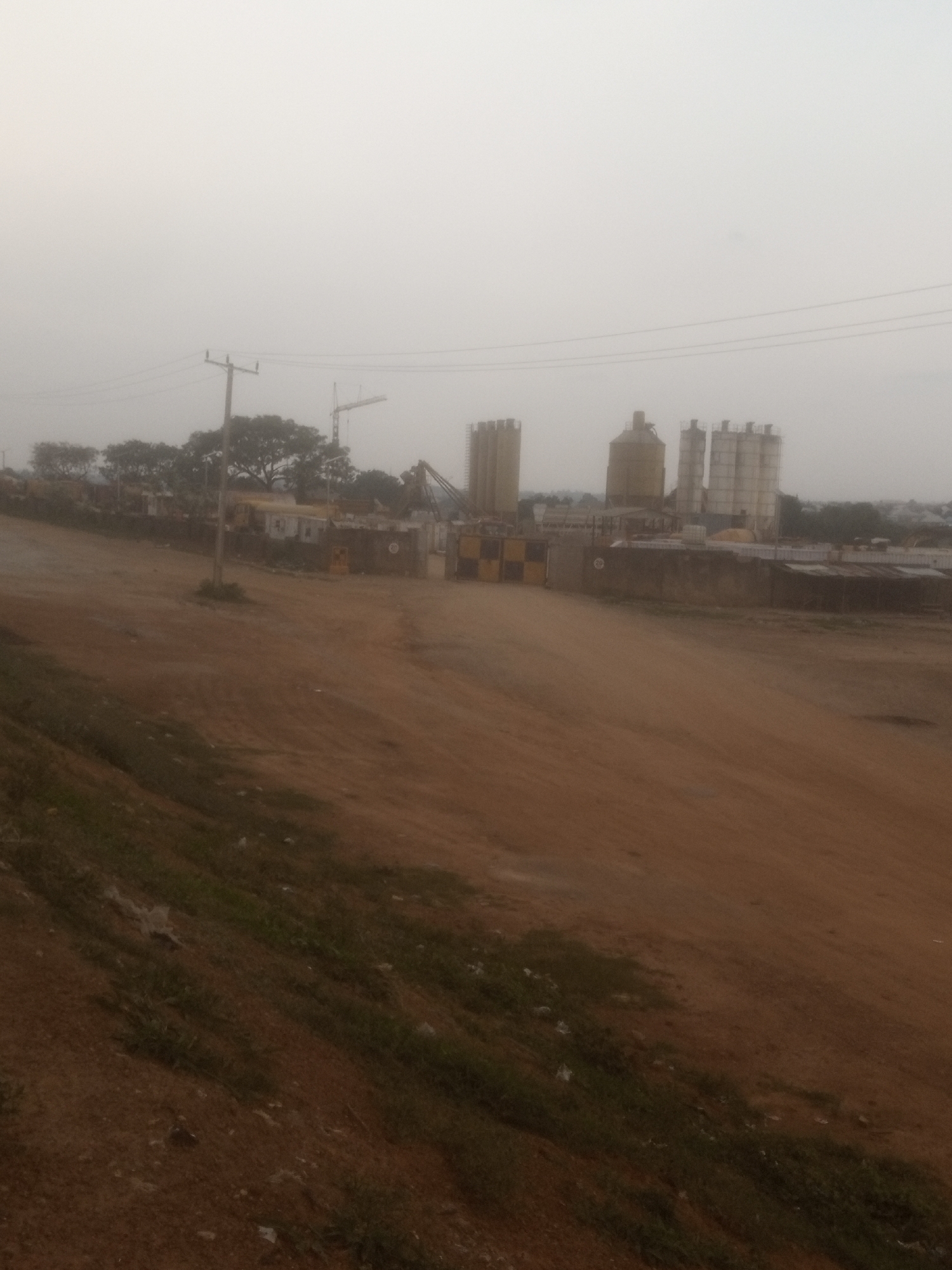 An outdoor, eye-level, medium shot captures a hazy industrial site in Sabon Gari, Nassarawa, Nigeria. The foreground is dominated by a dusty, reddish-brown dirt road that slopes down from left to right, with patches of sparse green grass growing on the embankment on the left. In the midground, a series of industrial structures rise against a pale, overcast sky. These include several tall, cylindrical silos, some appearing metallic and others concrete. To the left, a utility pole with wires is visible. Further left, trees with dense foliage are scattered. Several cranes, including a large tower crane, are present, suggesting ongoing construction or operation. Yellow and black checkered barriers are positioned near some of the structures. The background shows more buildings and greenery, blending into the misty atmosphere. The overall impression is one of an active industrial zone under a diffused, grey sky.