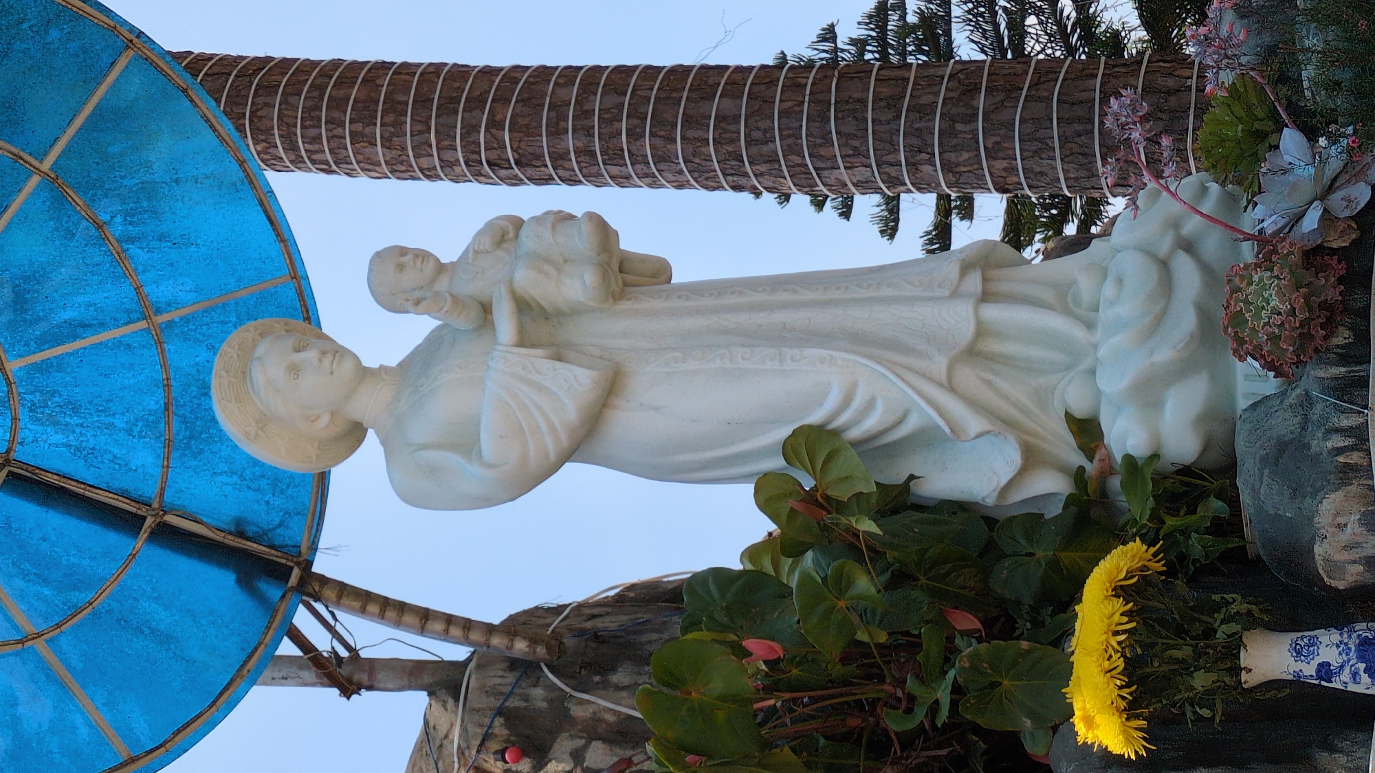 A statue of a woman holding a child is positioned outdoors in Đa Thọ, Vietnam. The statue, likely made of white stone or painted white, stands on a base adorned with green foliage, succulents, and a vase of yellow flowers. Behind the statue is a decorated tree trunk wrapped with rope and a blue, semi-circular structure. The sky is visible in the background.