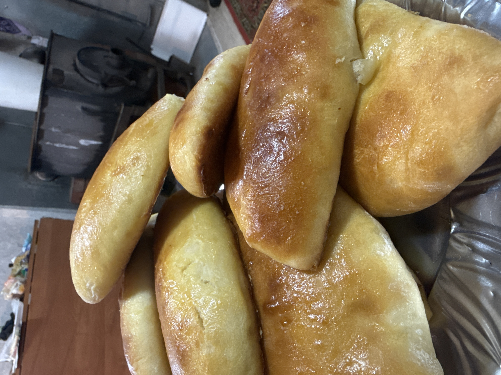 A close-up shot shows a pile of golden-brown pastries, likely buns or pies, glossy with a buttery or sugary glaze. They are arranged in an aluminum foil container, suggesting they might be fresh from the oven or intended for serving. The lighting is soft, emphasizing the texture and color of the baked goods. In the blurred background to the left, there is a dark, possibly metal, object which appears to be a stove or oven. No people are visible in the image. There is no definitive information in the photograph that confirms the location is in Chaikovsky, Russia, or any other place.