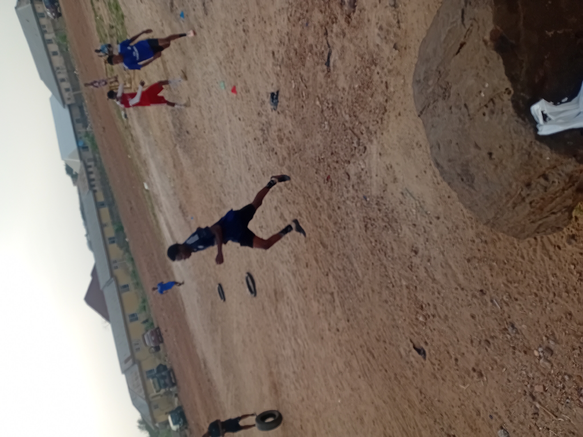 The image shows a sandy field in Unknown, Unknown, with several people engaged in what appears to be an athletic training session. Three individuals are visible running on the field, wearing athletic attire. There are also black tire-like objects scattered on the ground, presumably used as part of the training drills. In the background, there are buildings and parked cars, suggesting an urban setting. A large, circular indentation is visible in the foreground of the image.