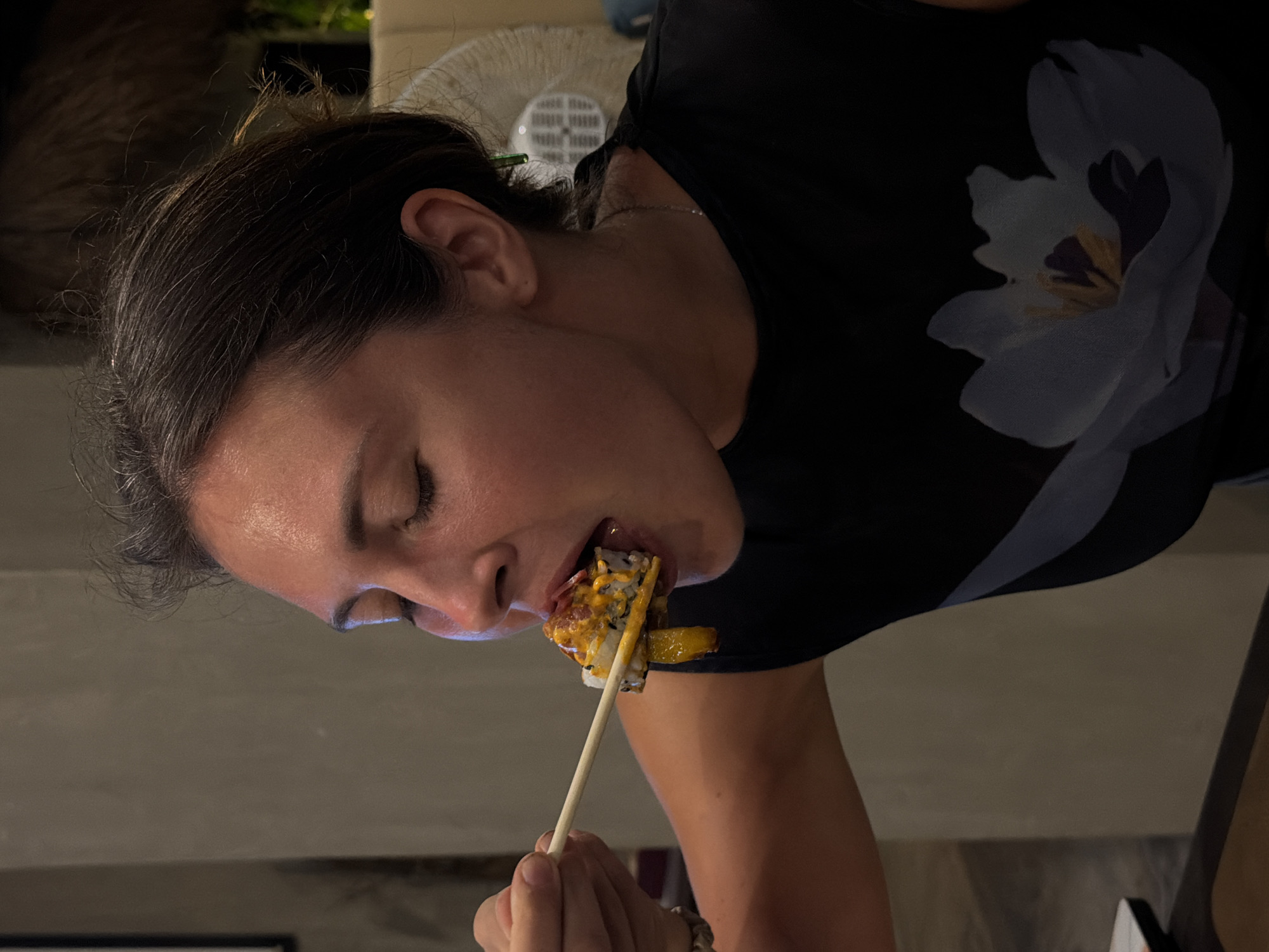 The image shows a close-up, profile view of a woman with dark hair eating sushi. Her eyes are closed, and she is in the process of taking a large sushi roll into her mouth using chopsticks. She is wearing a dark t-shirt featuring a prominent white and blue floral design on the shoulder area, and a thin chain necklace is visible around her neck. The sushi roll appears to have a creamy yellow topping and other ingredients. The scene is dimly lit, suggesting an evening or nighttime setting. The background is largely out of focus, revealing indistinct shapes including a white, possibly fan-like, object and some dark, blurry elements. The woman's expression conveys focus and enjoyment of her meal. No text is visible. The location is General Luna, Philippines.