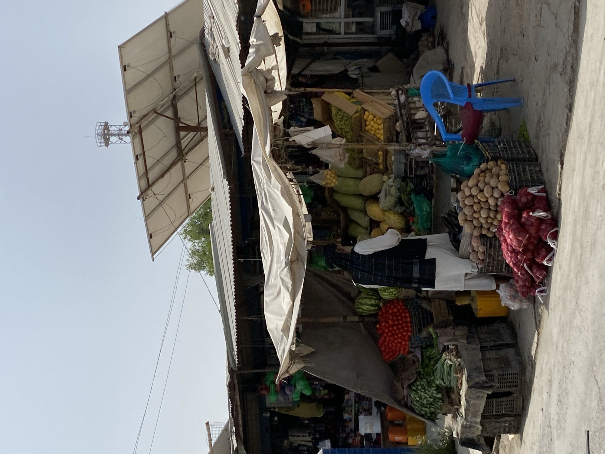 In the city of Kandahar, Afghanistan, an outdoor market stall is depicted under a clear sky. A person, dressed in traditional clothing with a dark checkered vest over a white garment and a dark headscarf, stands behind a display of fresh produce. The stall features an array of fruits and vegetables, including watermelons, tomatoes, potatoes, onions, green leafy vegetables, and various gourds. Some items are piled high in crates, while others are displayed in bags or on tables. A white tarp and corrugated metal roofing provide shade over the stall. In the background, a telecommunications tower is visible against the sky, and several solar panels are mounted on a structure above the market. A bright blue plastic chair sits in front of the stall. The ground is paved with concrete or similar material. The scene appears to be a typical day in a bustling market environment.