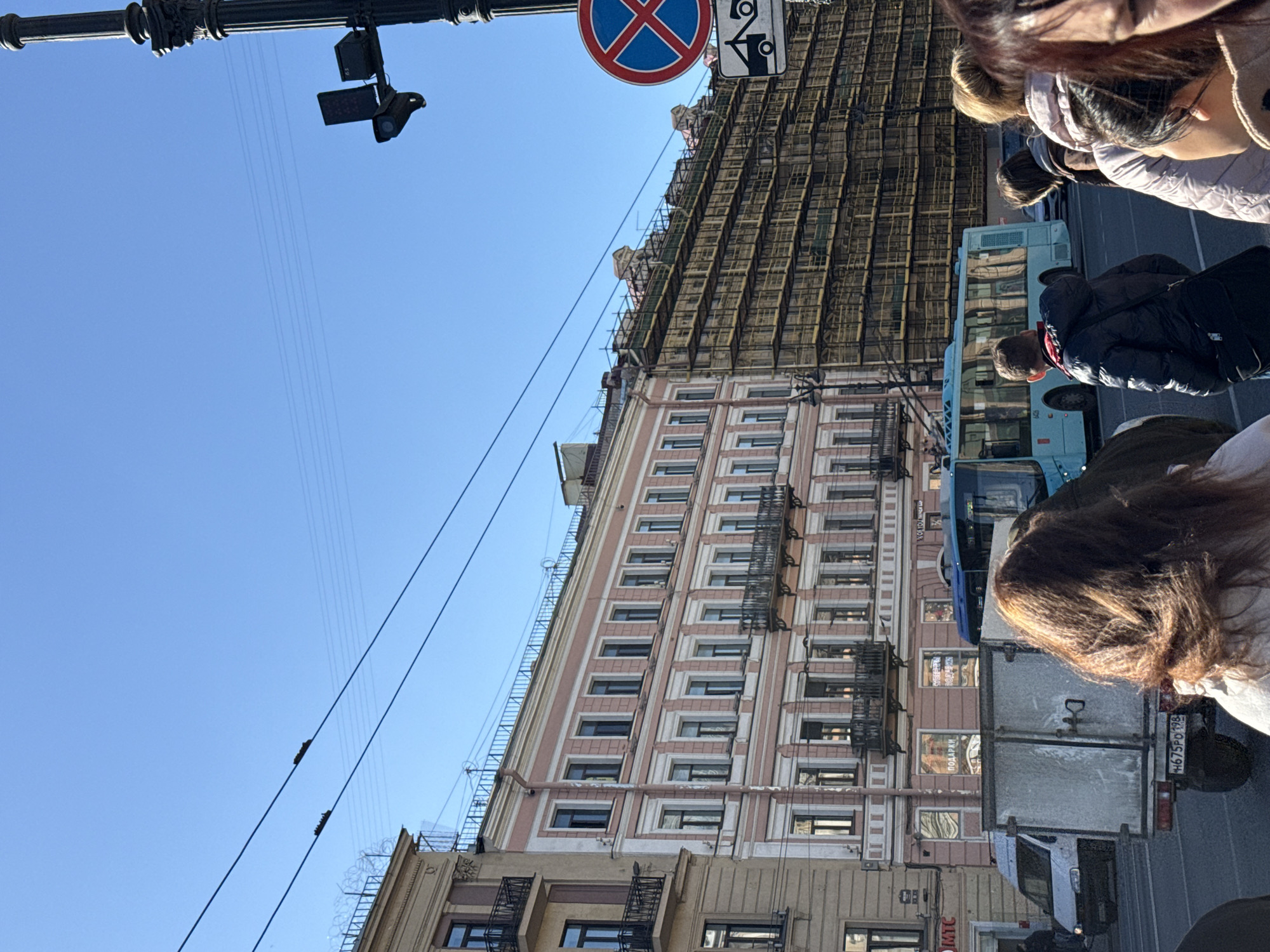 The image depicts a street scene in Saint Petersburg, Russia. A tall building with scaffolding covering its upper portion dominates the background. The building has a pink and white facade with numerous windows and fire escapes. A blue bus and a white delivery truck are parked on the street in front of the building. Pedestrians are present, partially obscuring the view. Overhead, there are traffic signals and a "No Parking" sign attached to a light post. Power lines stretch across the blue sky. The overall impression is of a bustling urban environment undergoing construction or renovation.